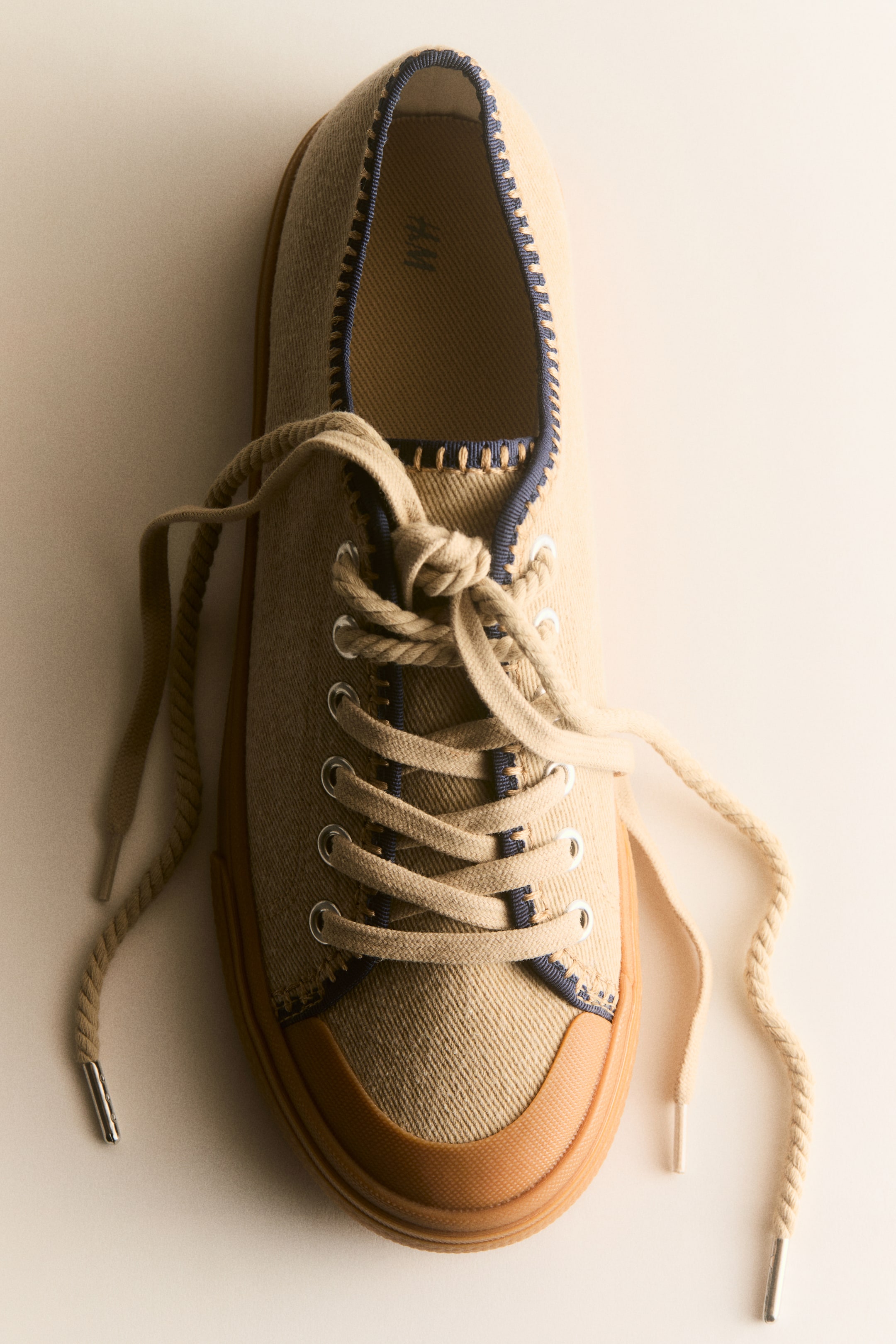 Größeres Bild anzeigen: Ein hellbrauner Canvas-Sneaker ist aus der Vogelperspektive nach vorne gerichtet und zeigt eine braune Gummikappe und -sohle. Er verfügt über silberne Ösen mit hellbraunen Schnürsenkeln, die zu einem Knoten gebunden sind, und dunkelblaue gewebte Verzierungen an Kragen und Schnürbereich mit hellen Nähten.