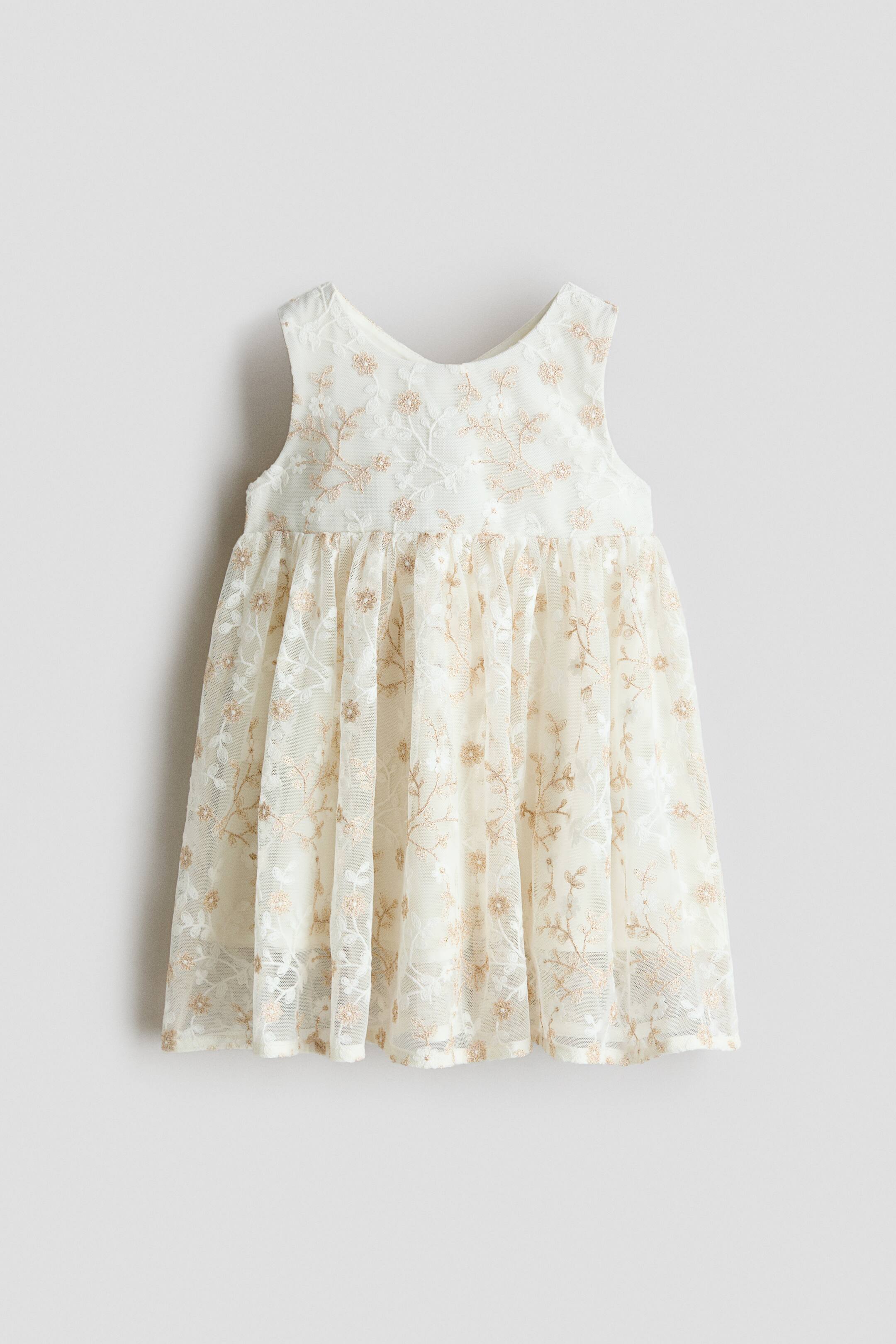 הצגת תמונה גדולה יותר: שמלת קרם ללא שרוולים מציגה שכבת רשת שקופה, מעוטרת ברקמת פרחים זהובים מתוחכמת וקישוטים קטנים ונוצצים, עם קו צוואר עגול, מותן מכווצת, וחצאית מלאה עם בטנה אטומה.