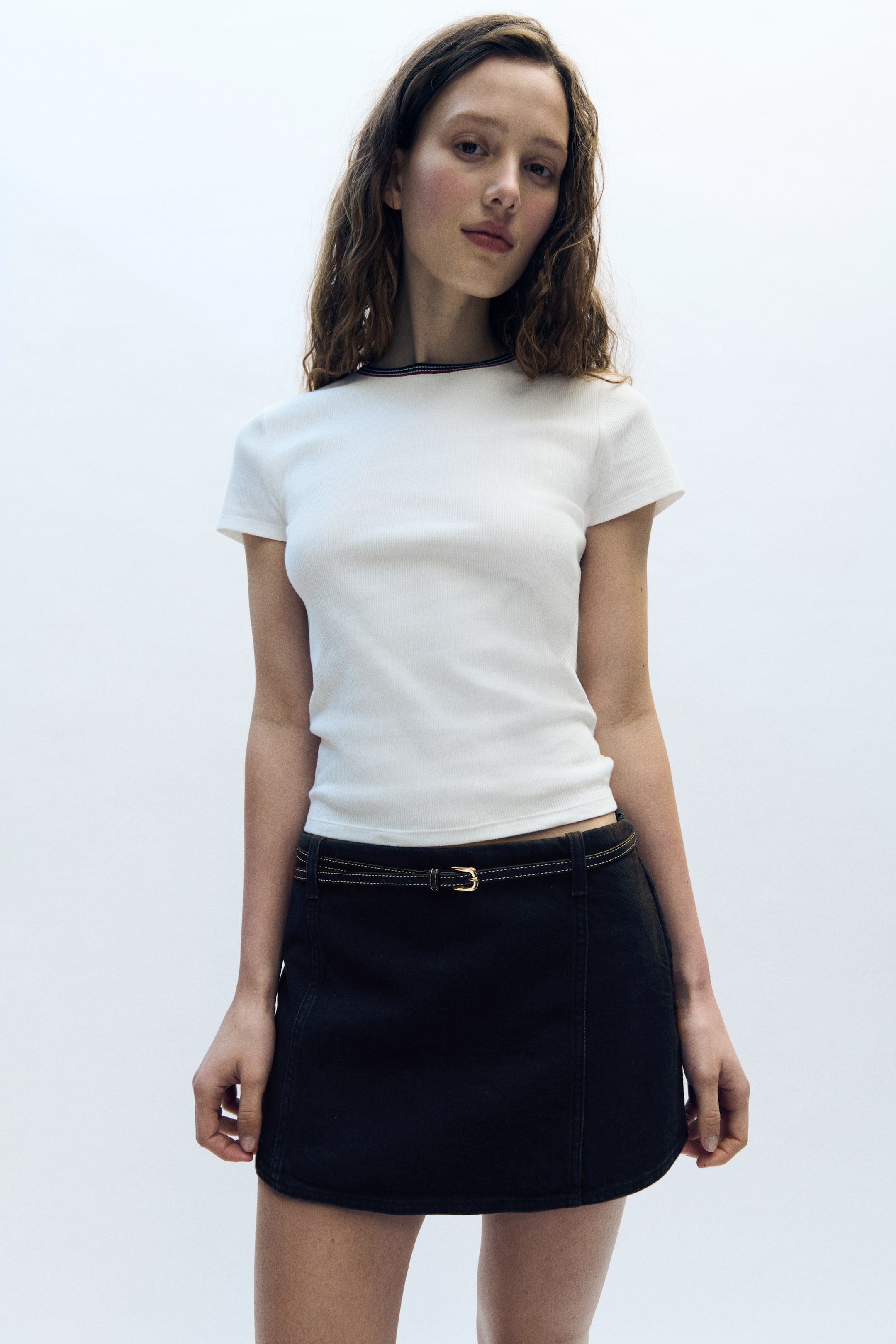 View larger image: A white ribbed short-sleeve t-shirt with a dark-trimmed crew neck is being worn, resting just above the waistband of a dark mini skirt. The mini skirt features visible stitching and is being styled with a thin dark belt with a gold buckle.