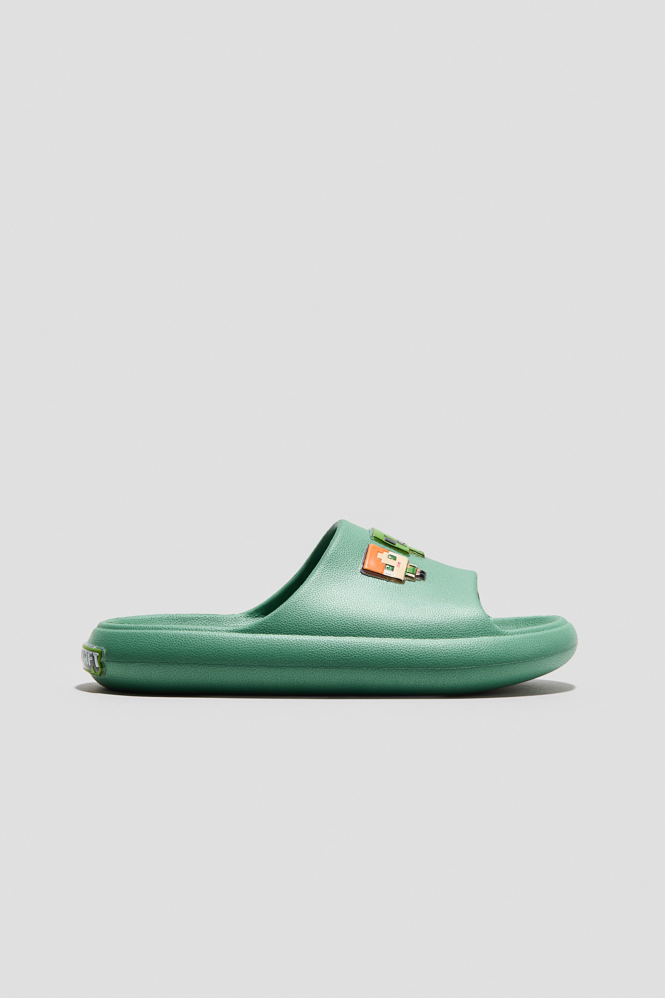 Ver imagen más grande: Una zapatilla de piscina verde se presenta de perfil lateral, con una correa ancha adornada con motivos pixelados, punta redonda, plantillas moldeadas y suelas gruesas.