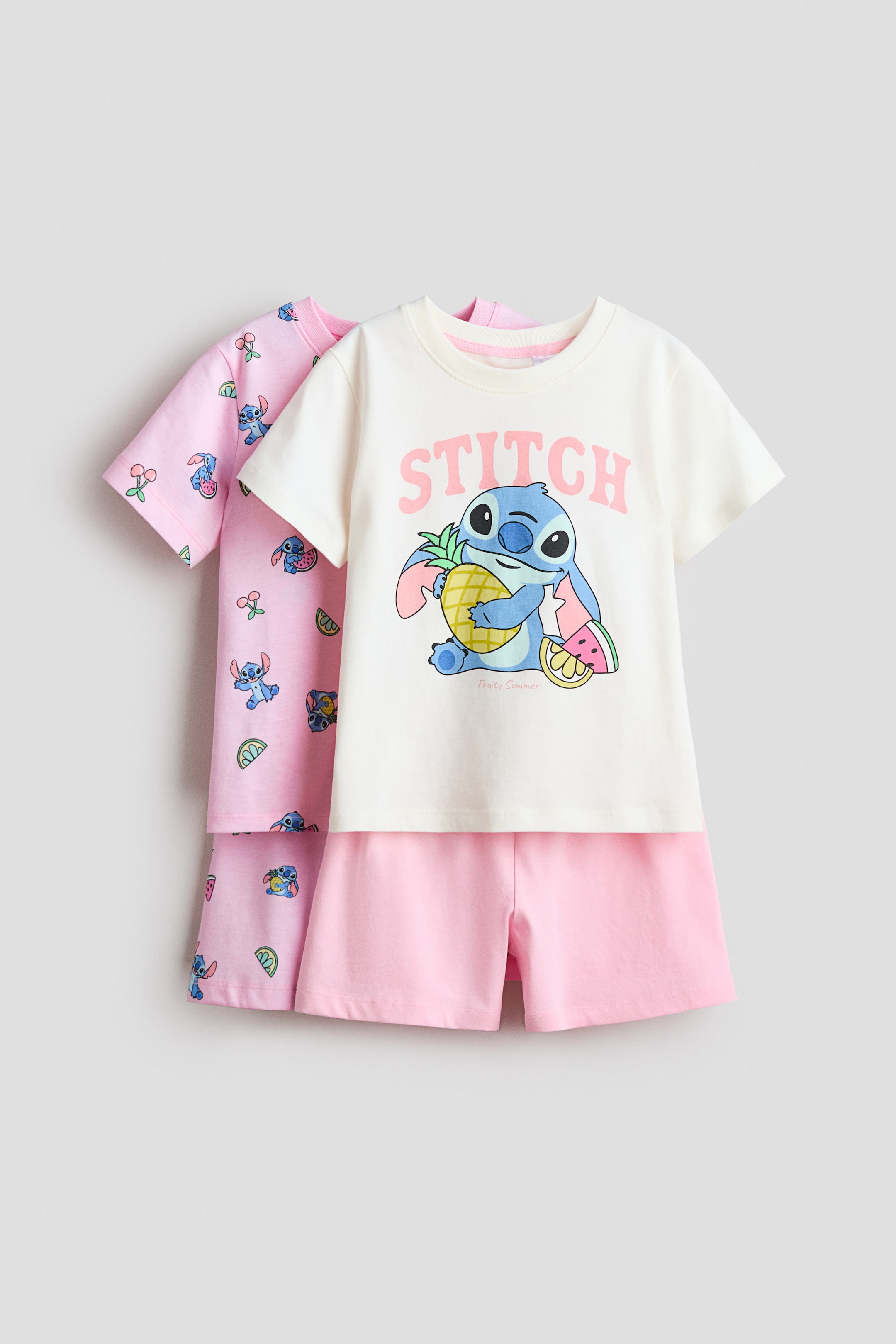 Grotere afbeelding bekijken: Twee sets korte mouwen pyjama's worden gepresenteerd, gelaagd achter elkaar, beide naar voren gericht. De voorste set bestaat uit een crèmekleurige top met een ronde halslijn en een grote grafische print van Stitch die een ananas vasthoudt, gecombineerd met effen lichtroze shorts. De achterste set, ook met een ronde halslijn en korte mouwen, is een lichtroze top en shorts met een all-over patroon van Stitch en fruitmotieven.