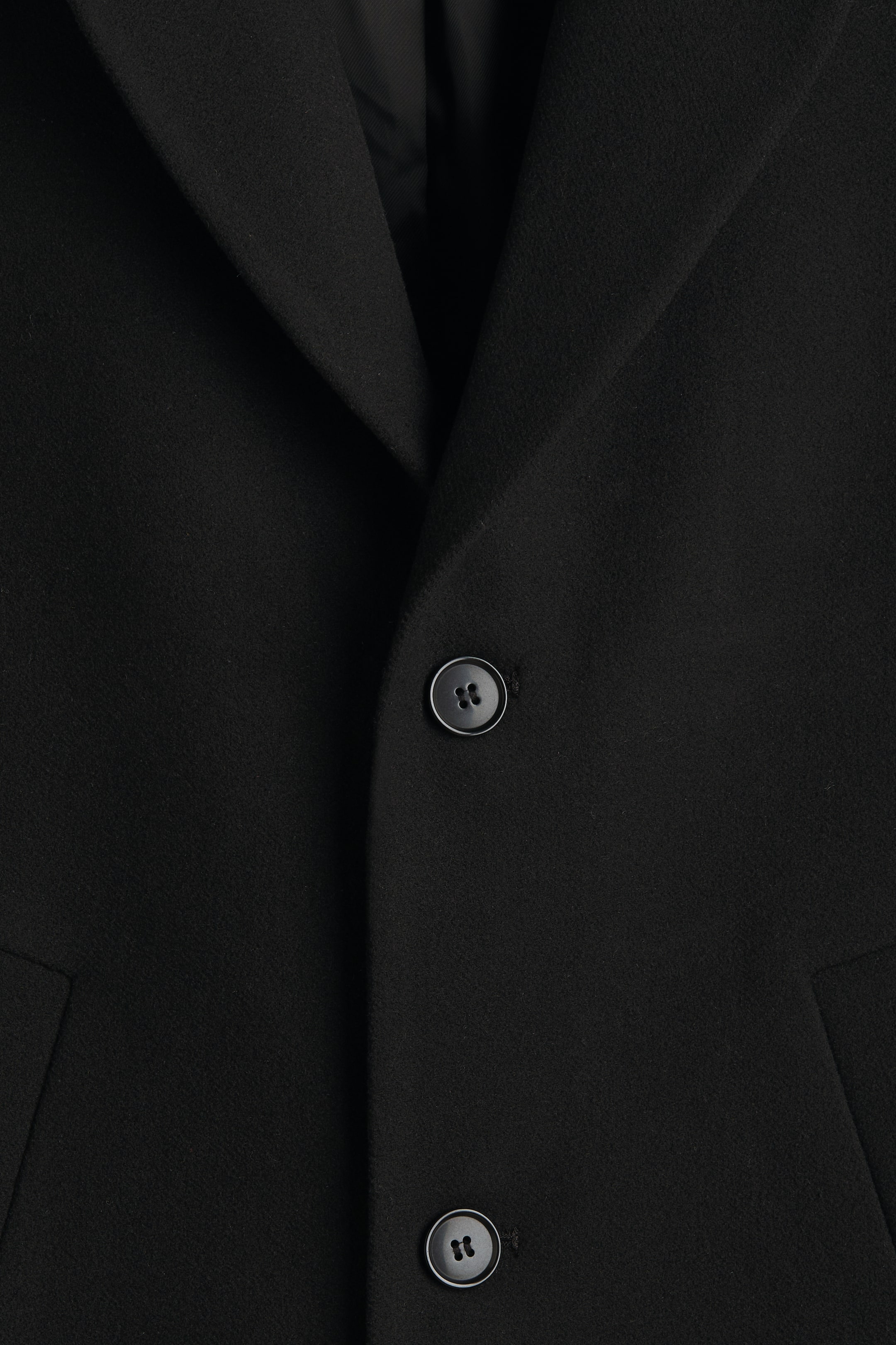 View larger image: A black coat is shown in a close-up view of its front panel, featuring a textured fabric. A folded lapel is visible on the left, and two circular black buttons, each with four holes, are positioned vertically on the right side, with a subtle pocket flap appearing below the lower button.