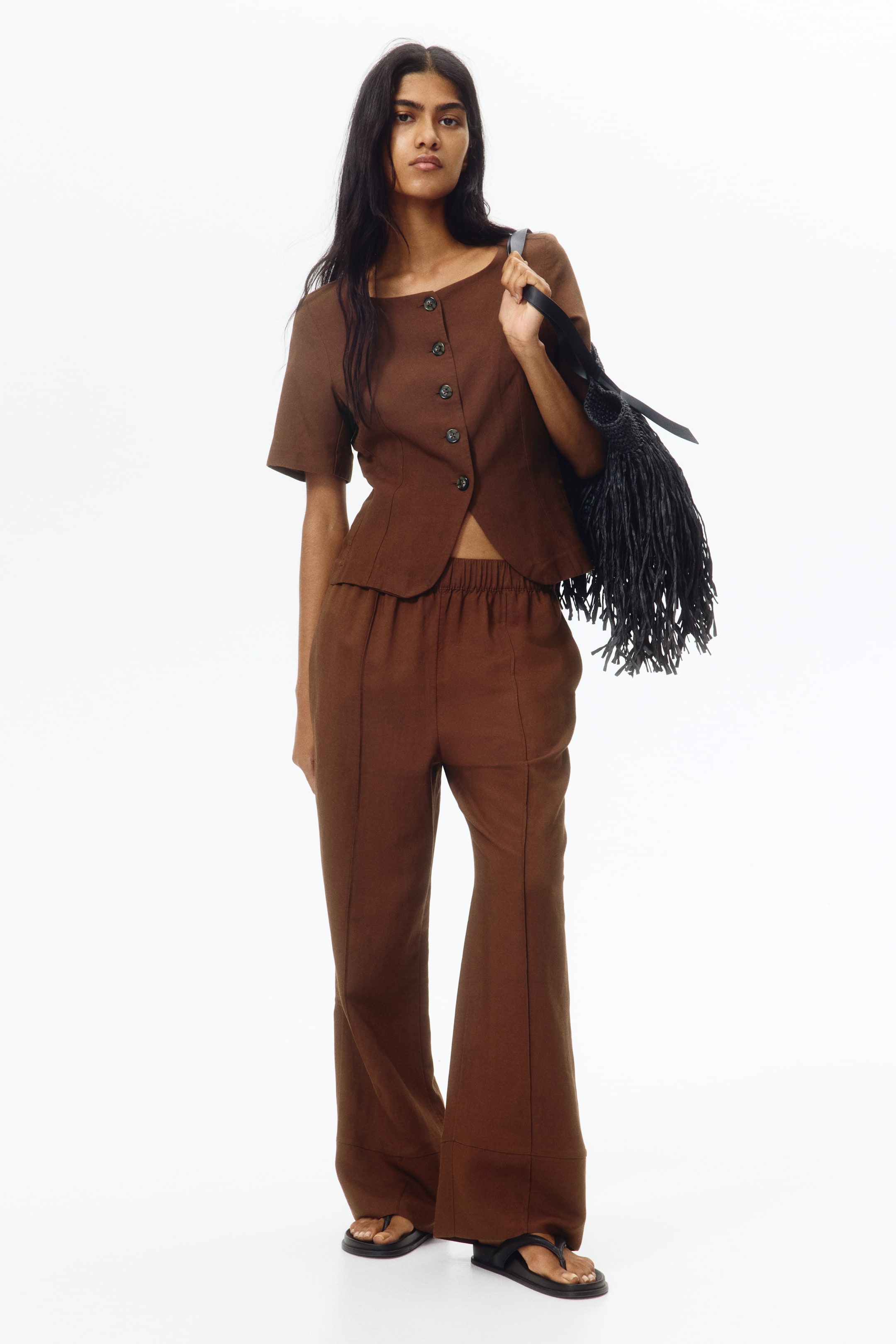 View larger image: A brown two-piece outfit crafted from a textured fabric showcases a short-sleeved top with a broad boat neckline, six front buttons, and a curved hem. It is paired with matching wide-leg pants featuring an elasticated waist and vertical front seams. Complementing the ensemble is a black fringed shoulder bag, and black thong sandals complete the look.