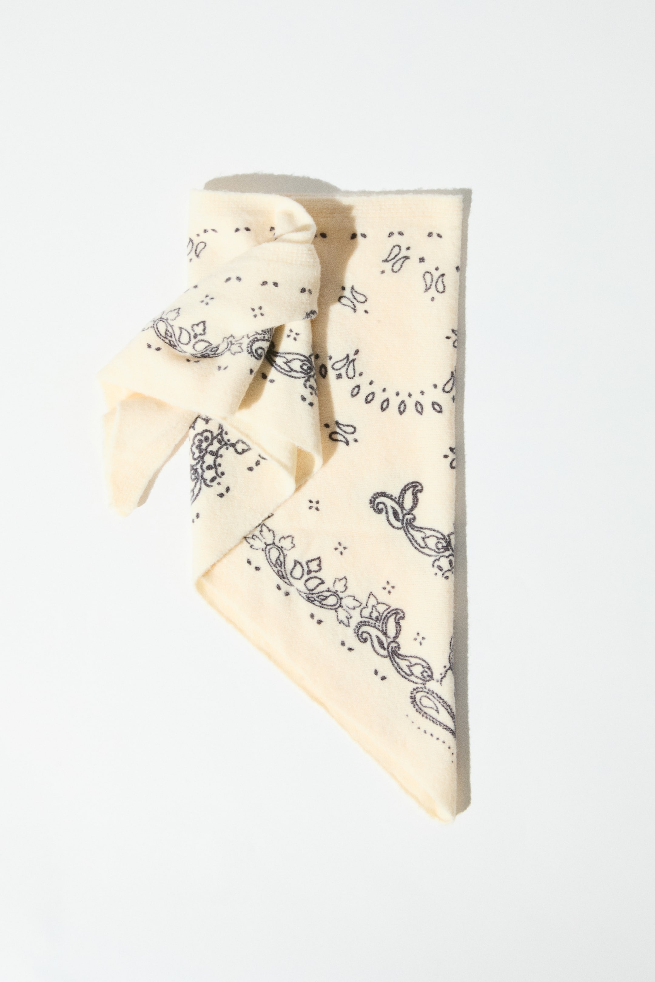 View larger image: A triangular scarf in a fine off-white knit is shown from the front with a printed dark paisley pattern. The scarf is folded flat with its pointed corner facing downward, and one end is loosely knotted on the upper left side.