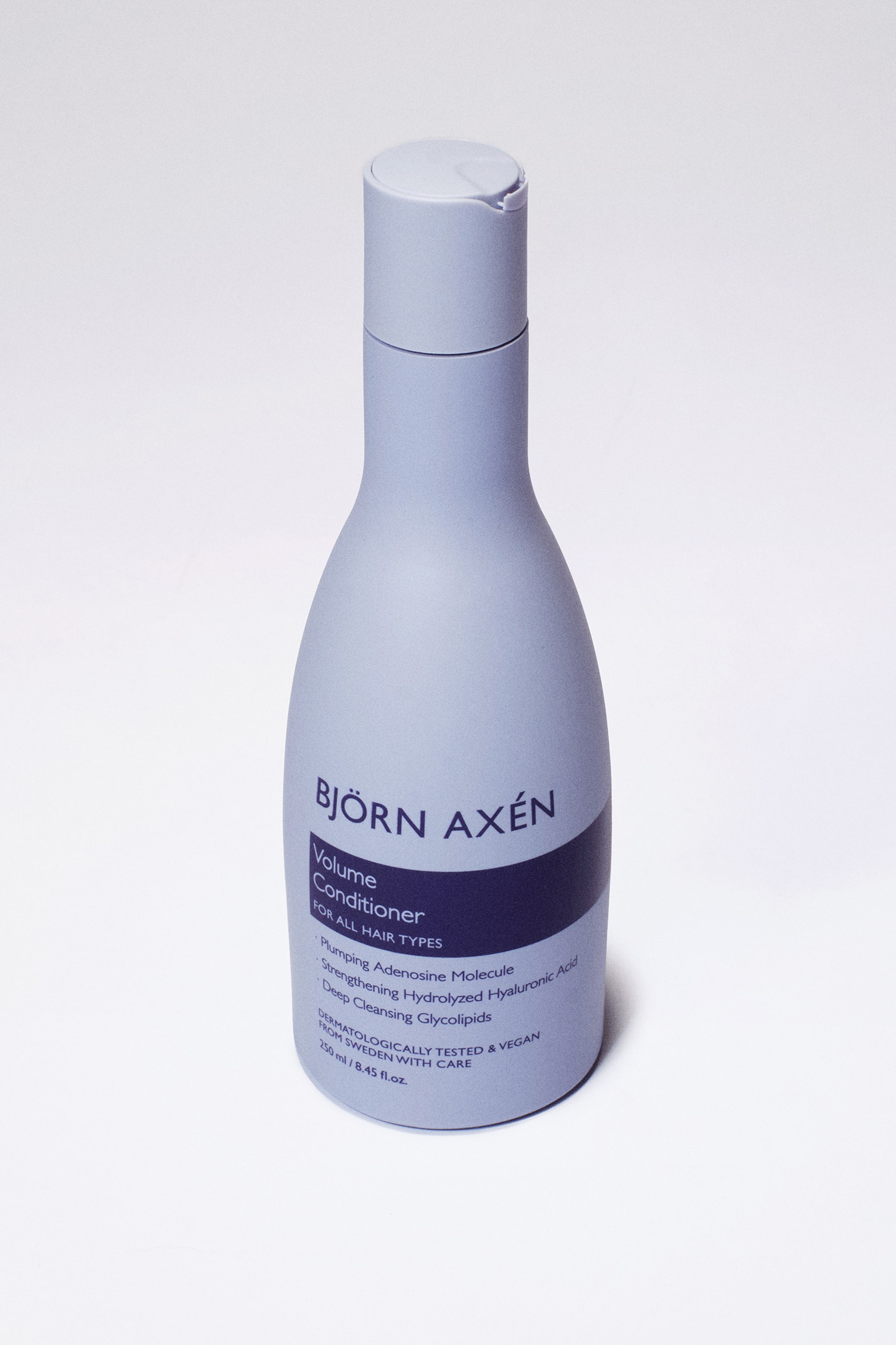 Se større billede: En dæmpet lavendel Björn Axén Volume Conditioner flaske står oprejst, let vinklet mod højre. Denne slanke, buede flaske har en mørk lilla-blå etiket med hvid tekst, der beskriver produktnavnet og nøgleingredienserne, og en matchende flip-top låg lukker toppen.