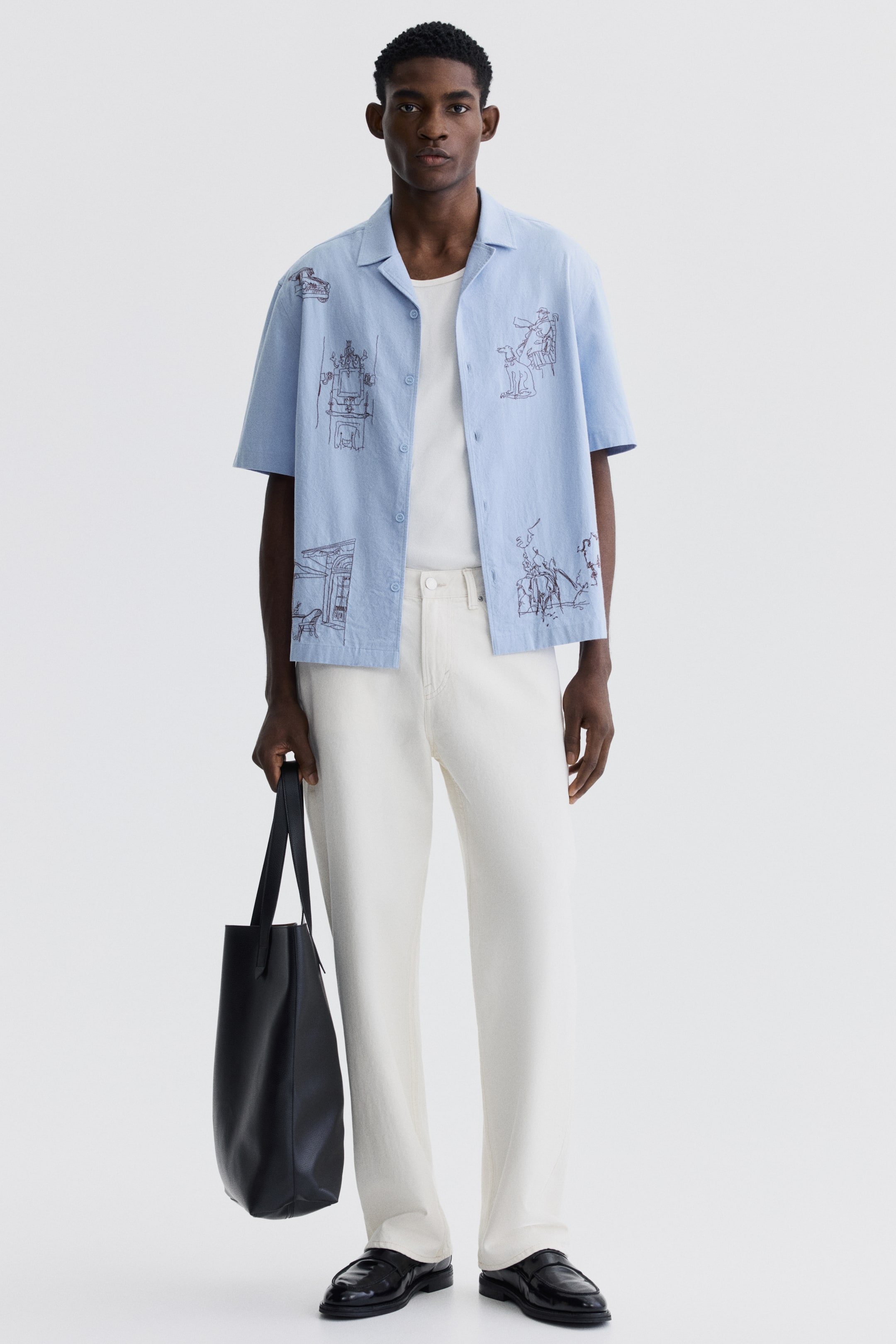 View larger image: A light blue short-sleeve shirt showcases dark brown line-art illustrations of buildings and figures. The shirt, featuring a revere collar and front button closure, is styled open over a white crew-neck t-shirt.