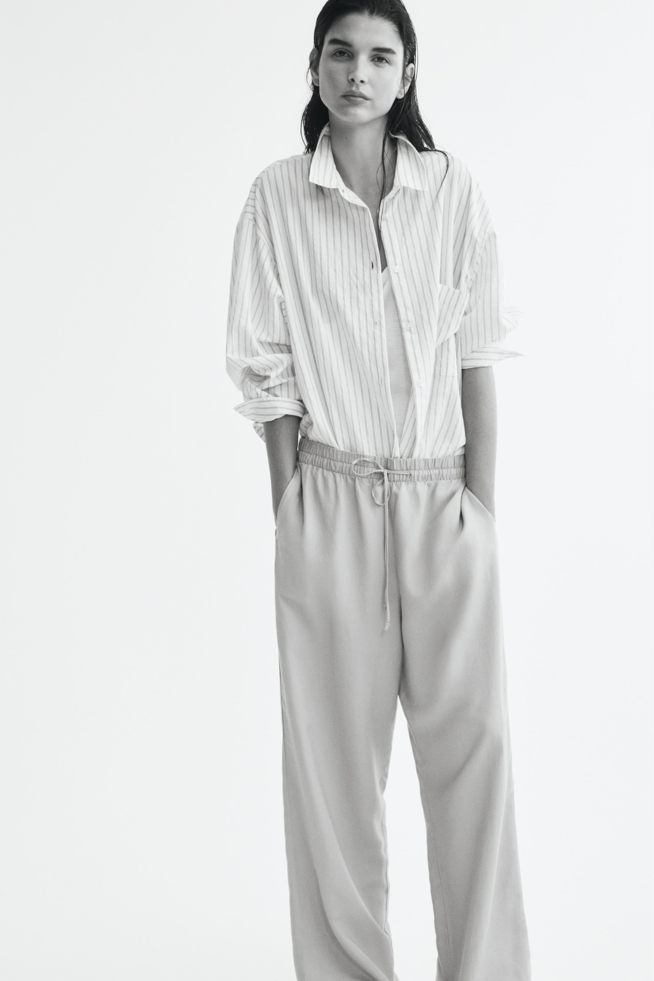 Agrandir l'image: Pantalon à coupe ample et jambe droite, doté d’une taille haute élastiquée avec un cordon de serrage étroit, porté par le mannequin, qui se tient debout avec les mains glissées dans les poches latérales. Le pantalon est associé à une chemise boutonnée à rayures partiellement rentrée.