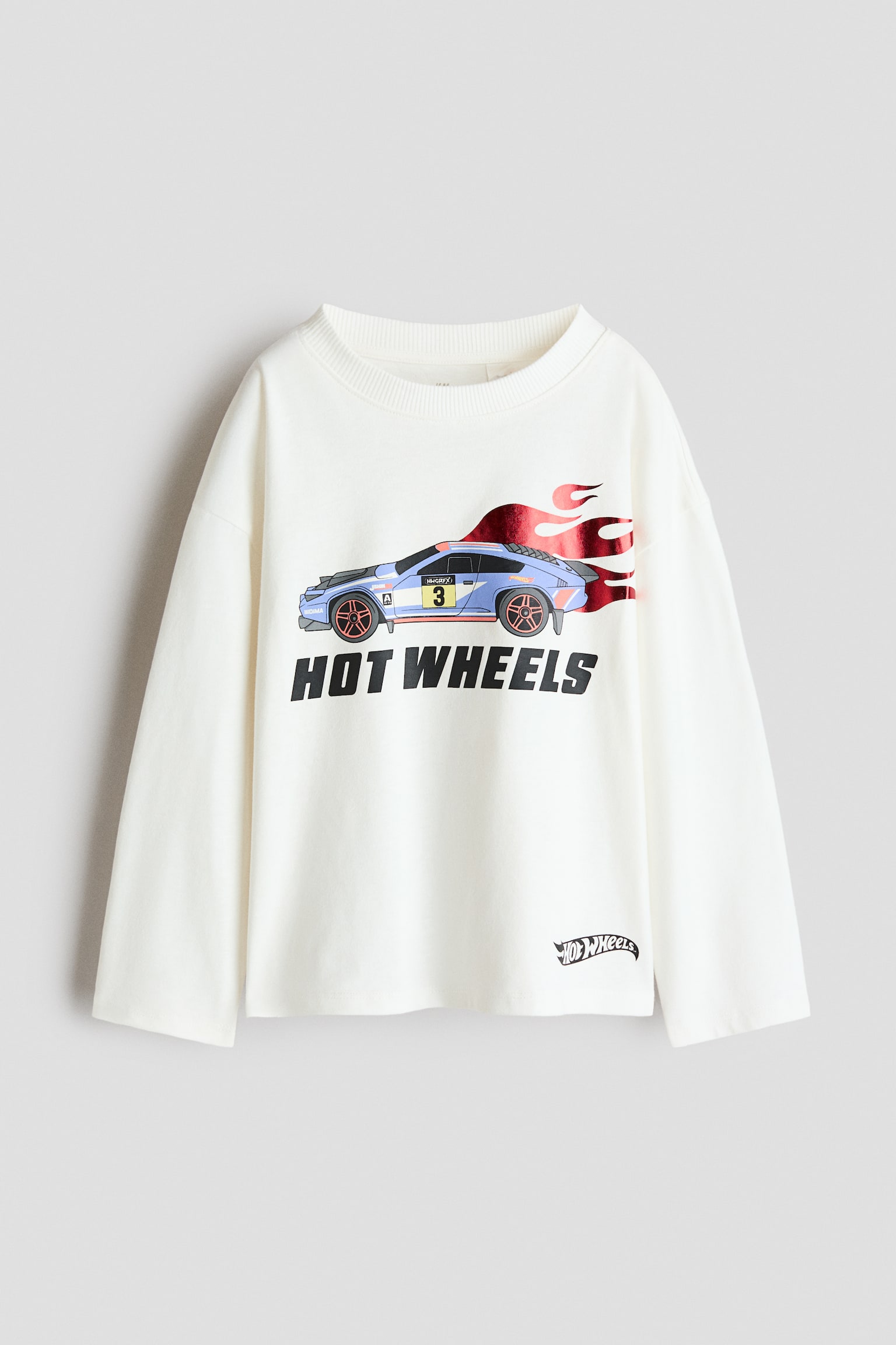 Vista frontale di una t-shirt a maniche lunghe, dalla vestibilità ampia, color bianco sporco, con scollo rotondo a costine, spalle scese e orlo dritto. Il davanti presenta una grafica di un'auto da corsa blu con scintillanti fiamme in lamina rossa che si estendono sopra il testo nero in grassetto "HOT WHEELS", con un logo più piccolo vicino all'orlo inferiore sinistro.
