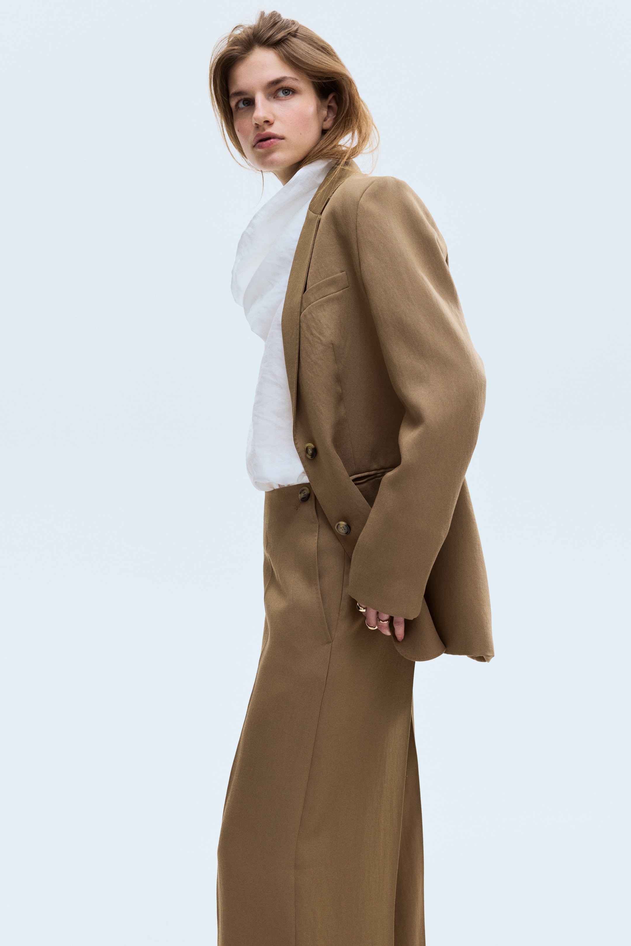 Ver imagen más grande: Un traje de dos piezas en color beige, compuesto por una chaqueta cruzada y pantalones de pierna ancha, se presenta desde un ángulo frontal. La chaqueta muestra solapas de muesca, dos botones oscuros y un bolsillo con solapa, mientras se lleva sobre una camisa blanca, y los pantalones presentan un cierre de botón y gancho en la cintura.