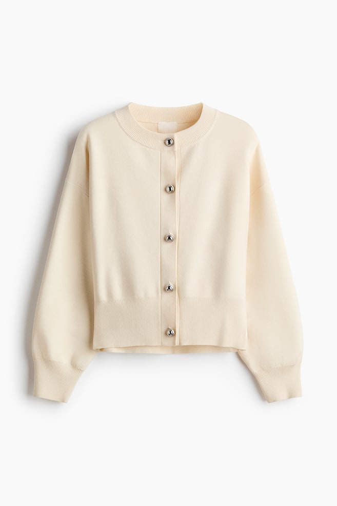A cream-colored, fine-knit cardigan is shown facing front, featuring a round, rib-trimmed neckline, dropped shoulders, and five silver-tone dome buttons down the center front. The cardigan is displaying long sleeves with wide ribbed cuffs and a matching ribbed hem. - 1