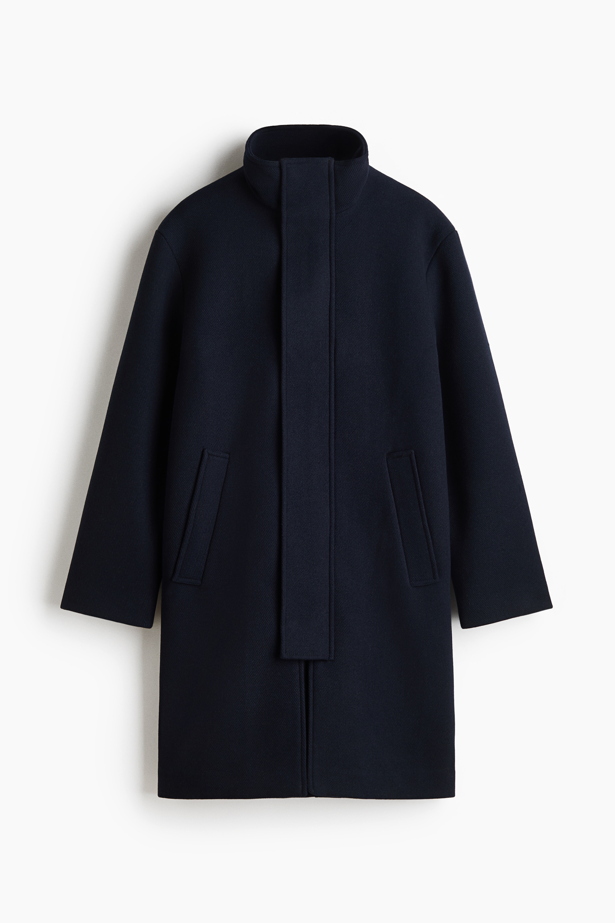 The model is wearing a knee-length dark navy felted twill coat featuring a high funnel collar, a concealed front zip closure with a wind flap, and side welt pockets. The coat is styled with dark trousers with side stripes, and the model is carrying a black bag under one arm.