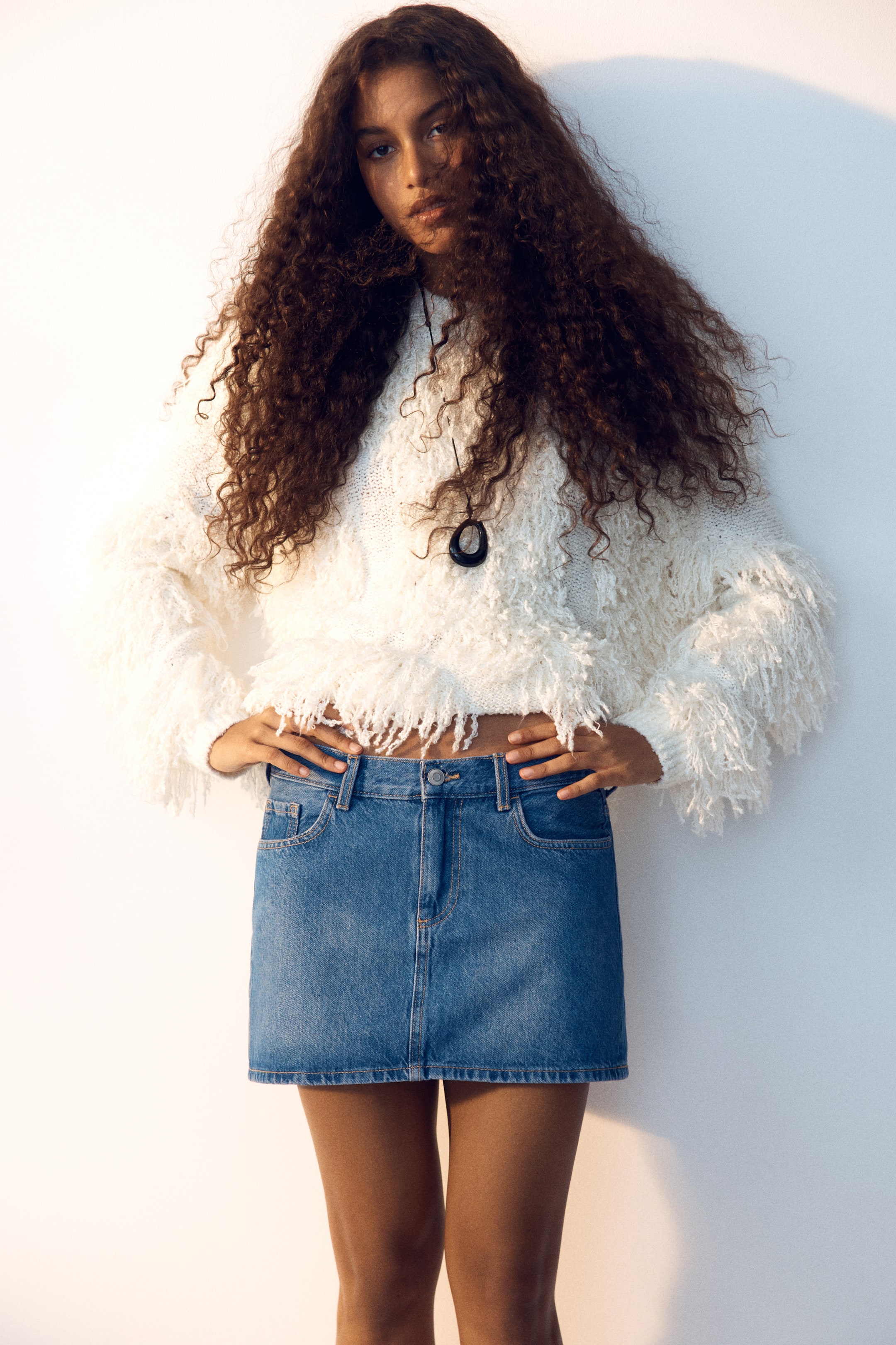 View larger image: The denim mini skirt showcases a medium blue wash, positioned directly forward. It features front pockets, belt loops, a button and zip fastening at the waist, and a straight hem, with its upper edge visible beneath a white cropped jumper.