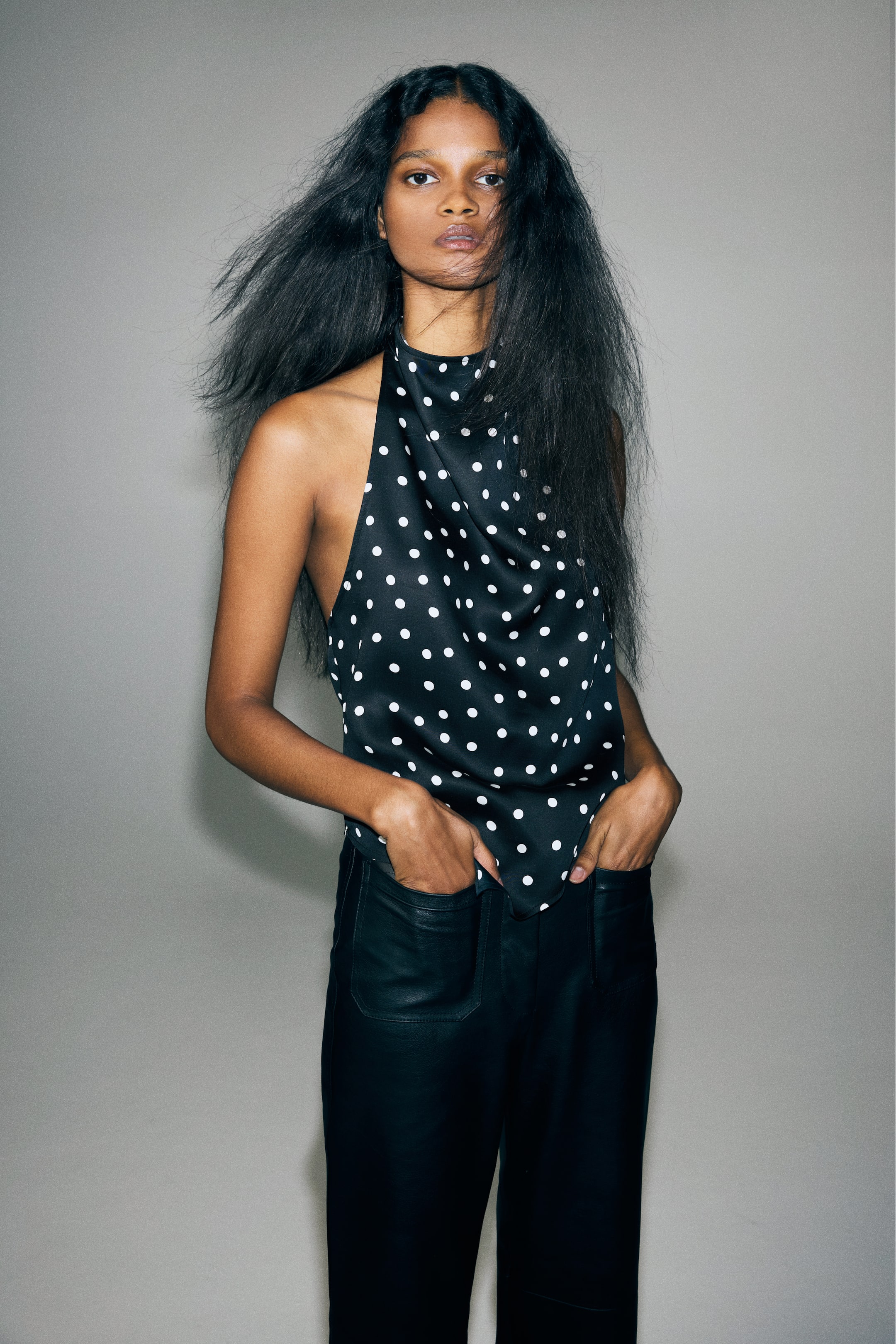 Grotere afbeelding bekijken: Een zwart satijnen haltertop met witte stippen wordt gedragen door het model, met een gedrapeerde watervalhals en een asymmetrische puntige zoom. De top is gestyled met een zwarte broek, waarin het model beide handen steekt.