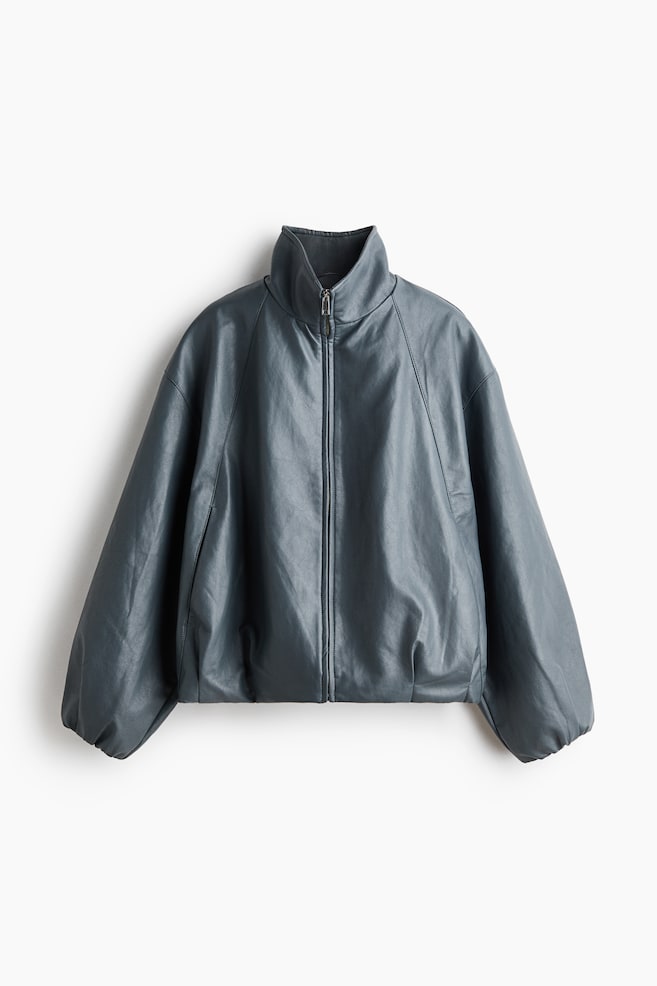 A muted blue-green jacket is showcased from the front, featuring a high stand-up collar and a central zip fastening. It is crafted with voluminous raglan sleeves and a gathered hem, creating a blouson silhouette. - 1