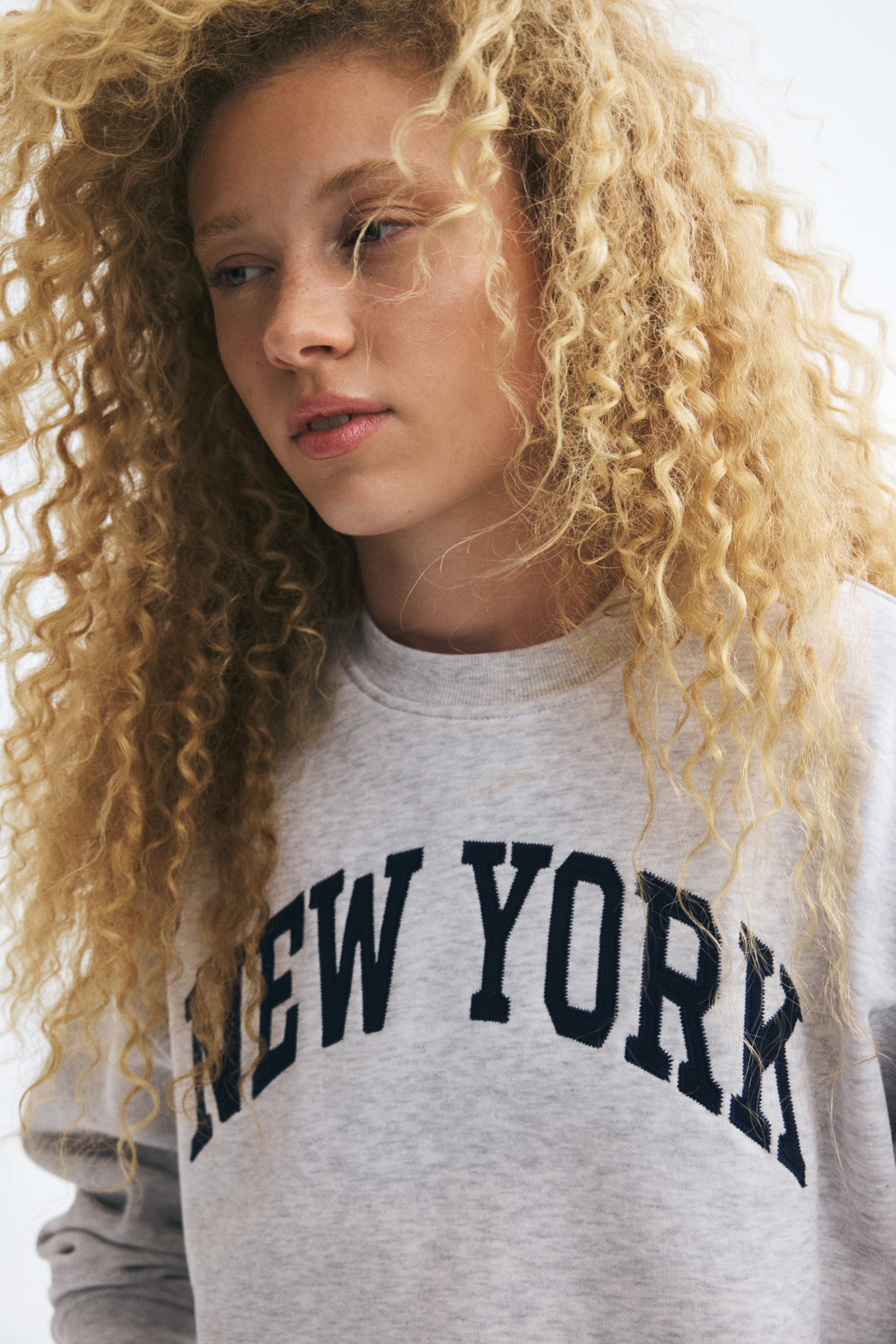 View larger image: A light grey sweat-fabric sweater featuring a round, rib-trimmed neckline and a dark blue "NEW YORK" printed design across the chest is being worn by the model. The sweater has a relaxed fit with dropped shoulders.