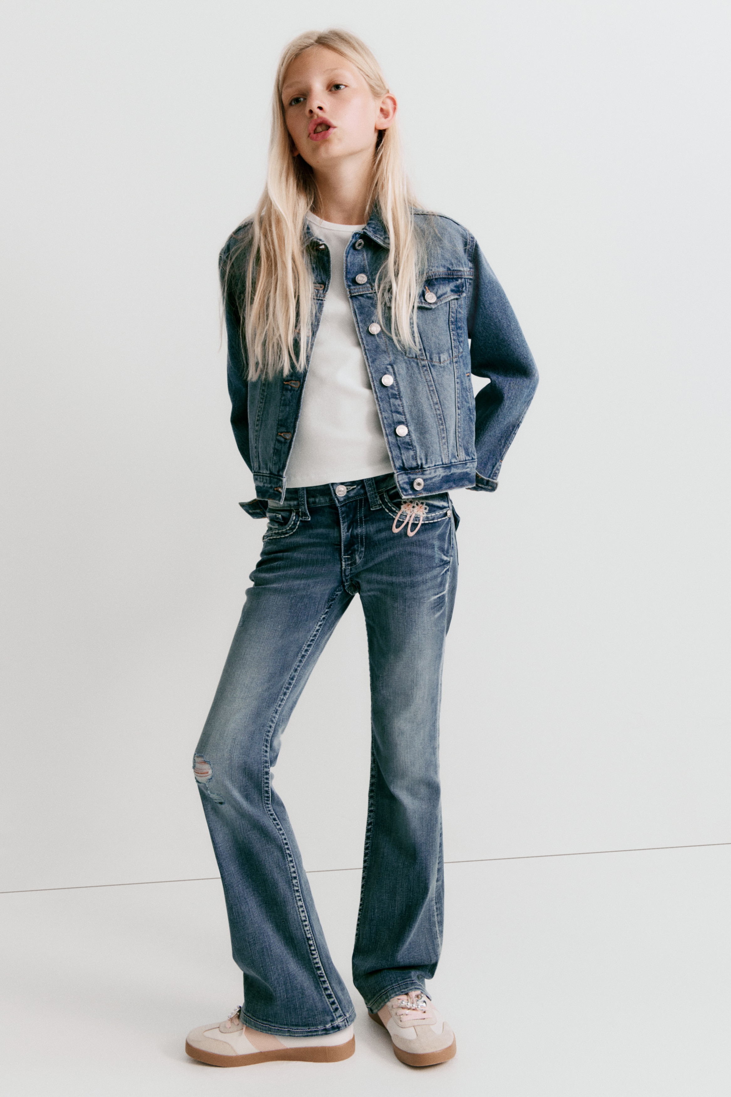 Jeans in denim lavato presentano una vista frontale, con vita bassa dotata di chiusura con zip e bottone, insieme a tasche anteriori e una tasca per monete. Mostrano dettagli consumati, tra cui baffature sulle cosce e una toppa rovinata sulla gamba sinistra, e sono progettati con una vestibilità skinny dalla vita alla coscia, allargandosi in una gamba a zampa dal ginocchio all'orlo.