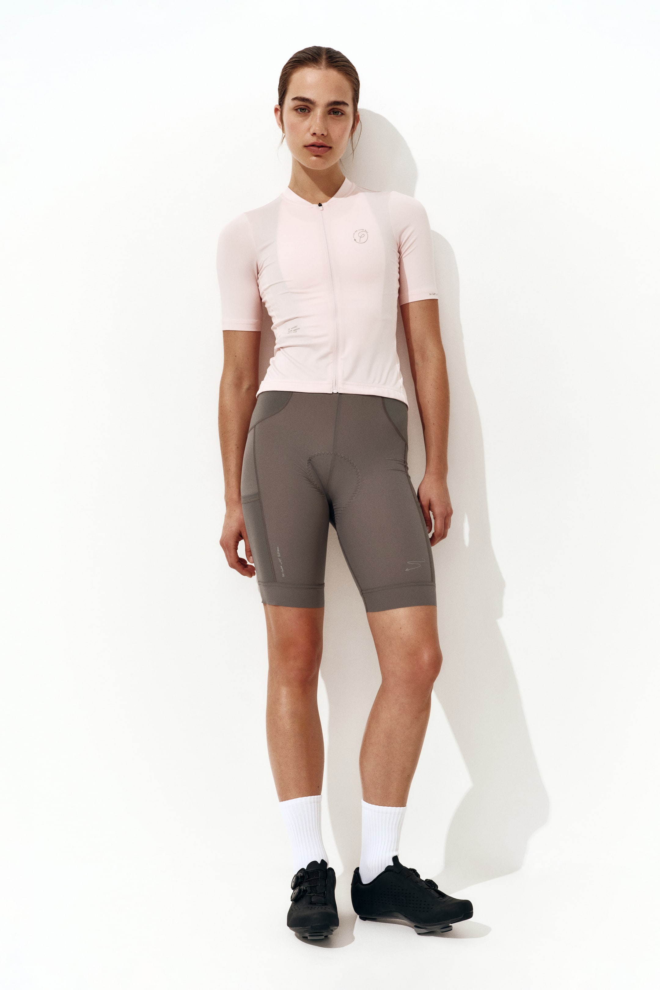 View larger image: A light pink short-sleeve cycling jersey, featuring a high collar, a full-length front zipper, and a circular chest logo, is shown from a front view. It is paired with dark gray cycling shorts that include thigh pockets and visible chamois padding, with both garments fitting snugly to the body.