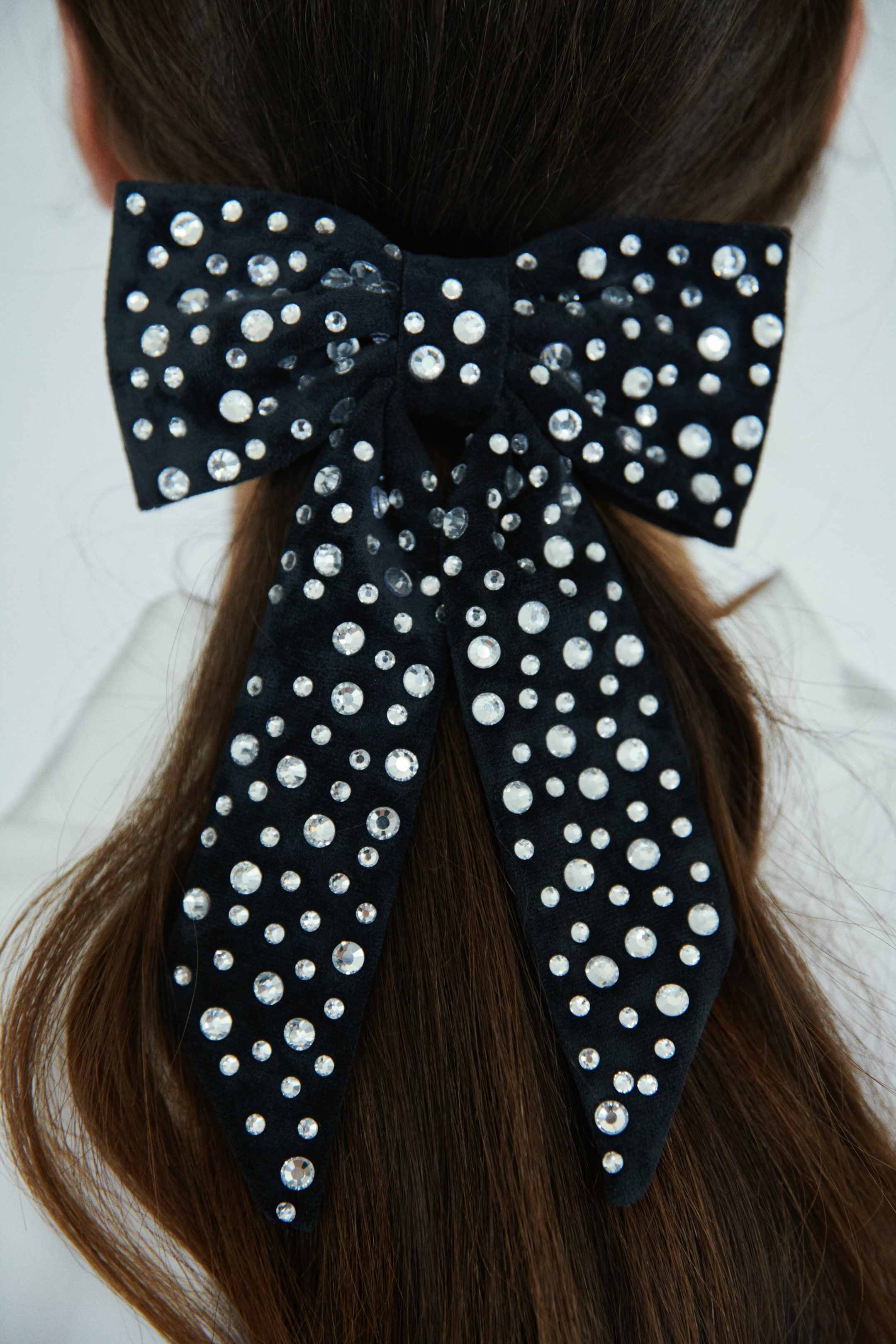 View larger image: A black velvet hair bow is being worn in brown hair, positioned at the back of the head. The bow is adorned with numerous clear, round rhinestones of various sizes, which are scattered across its surface and trailing ends.