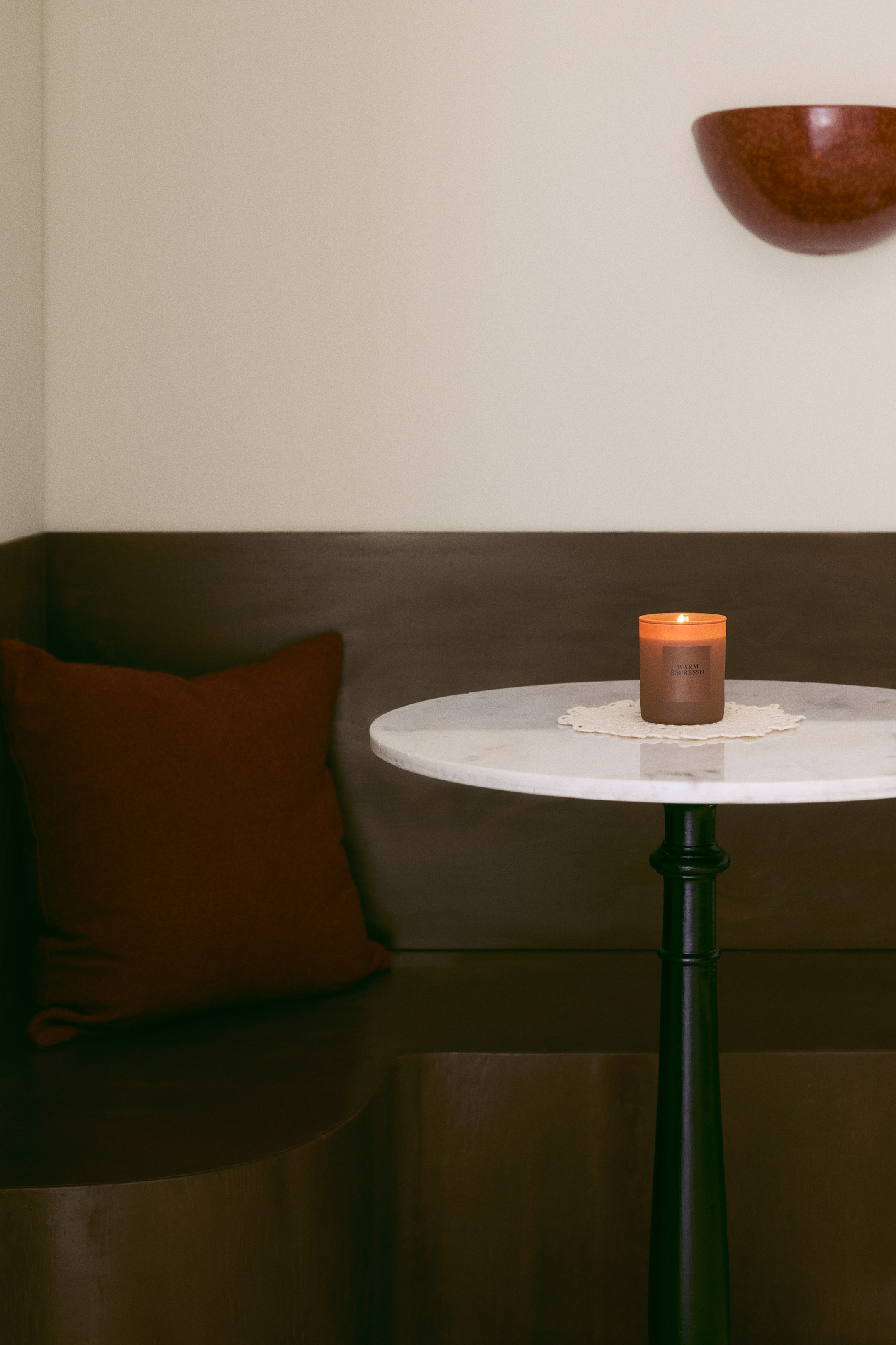 Voir une image plus grande: Une bougie parfumée dans un support cylindrique en verre brun étiqueté « WARM ESPRESSO » est allumée avec une petite flamme. La bougie est orientée vers l’avant et repose sur un petit napperon en dentelle blanche, sur un dessus de table en marbre.