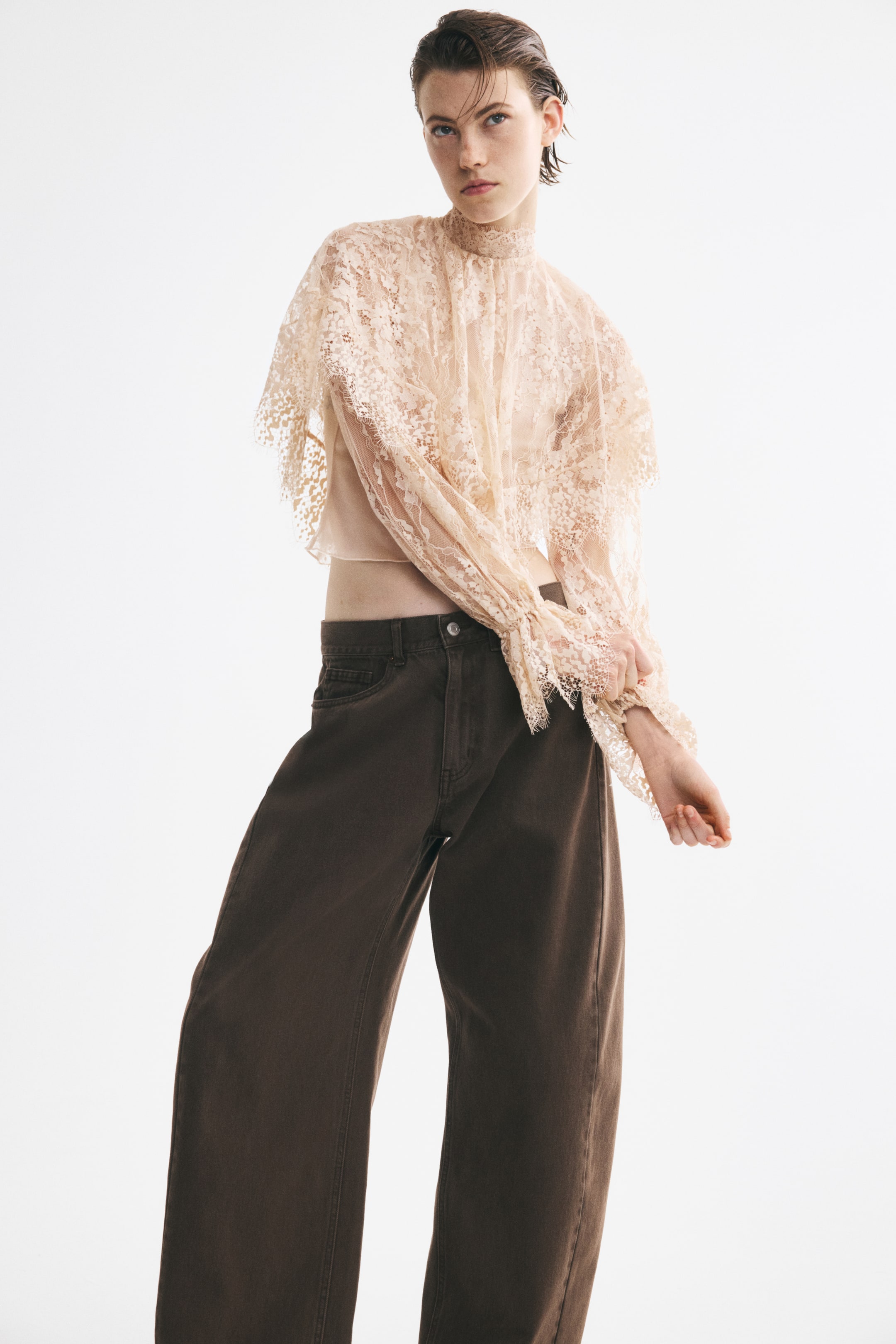 View larger image: A beige cropped lace top is shown from the front, featuring a high scalloped ruffle collar and a cape-like layer with a scalloped hem draping over the shoulders. The design showcases long balloon sleeves with narrow elasticized wrists and flounced, scalloped cuffs, highlighting a cropped fit styled with dark brown wide-leg trousers.