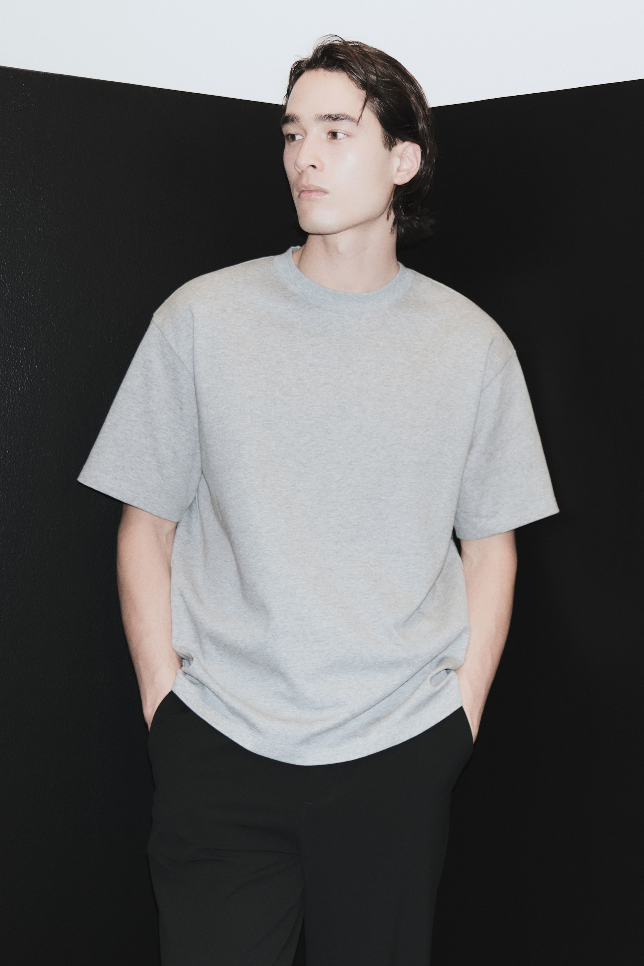 Xem ảnh lớn hơn: Chiếc áo thun cổ tròn màu xám nhạt được trình bày ở góc nghiêng trước bên phải, thể hiện dáng rộng (oversized) thoải mái. Áo có tay ngắn rộng và rũ nhẹ với gấu thẳng, dài phủ qua eo.