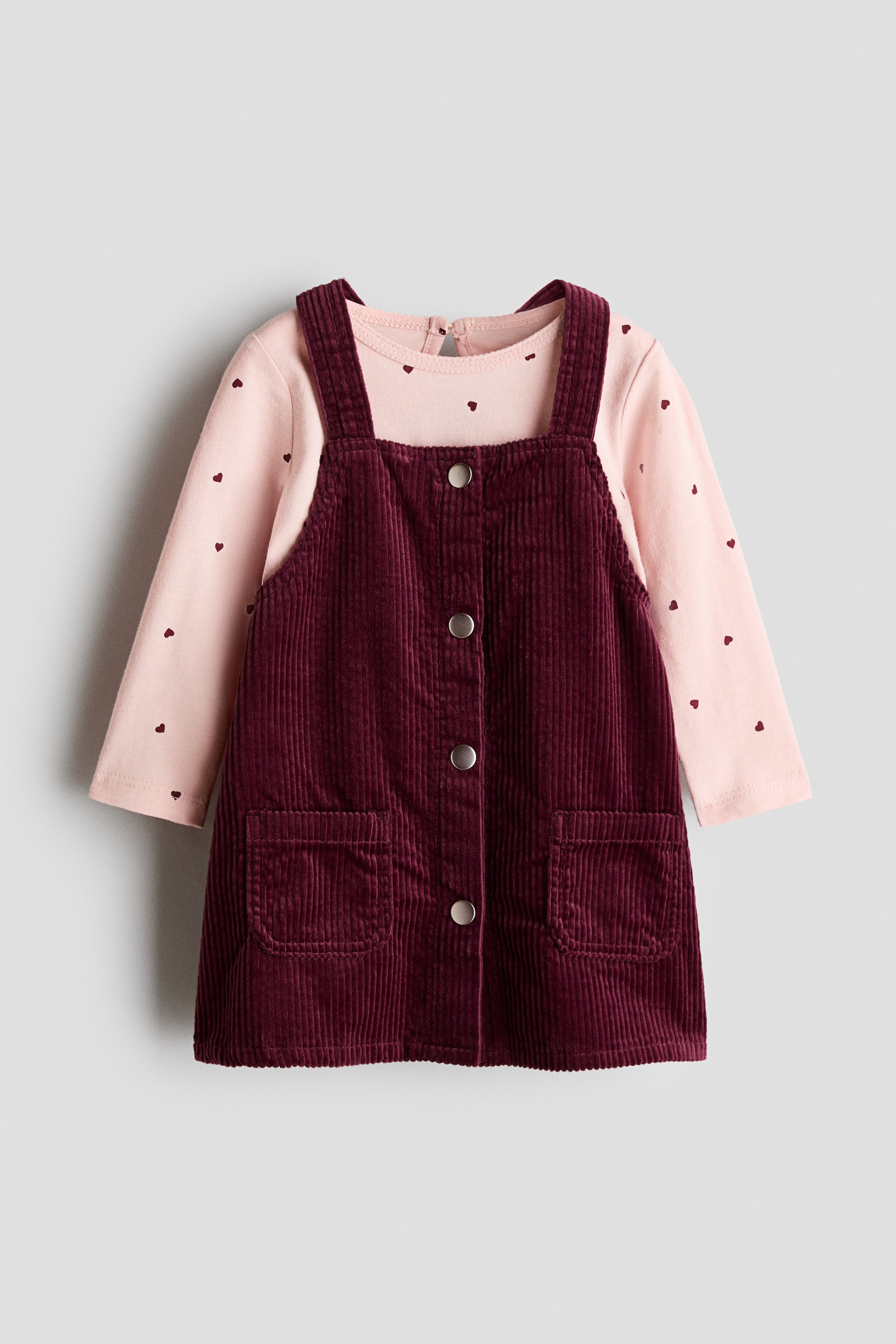 View larger image: A baby's outfit showcases a burgundy corduroy pinafore dress layered over a light pink long-sleeved top. The pinafore features shoulder straps, four silver-toned buttons down the front, and two square patch pockets. The pink top displays a pattern of small, scattered heart shapes and a round neckline with a back button closure.