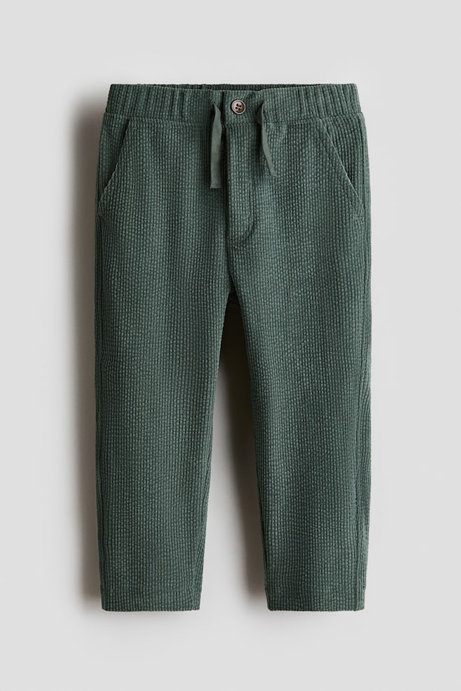 A pair of dark green, textured jersey trousers is hanging in a front-facing view, showcasing an elasticated waistband with a matching woven drawstring and a single brown button on a mock fly. The trousers feature a vertical ribbed pattern and diagonal side pockets. - 1
