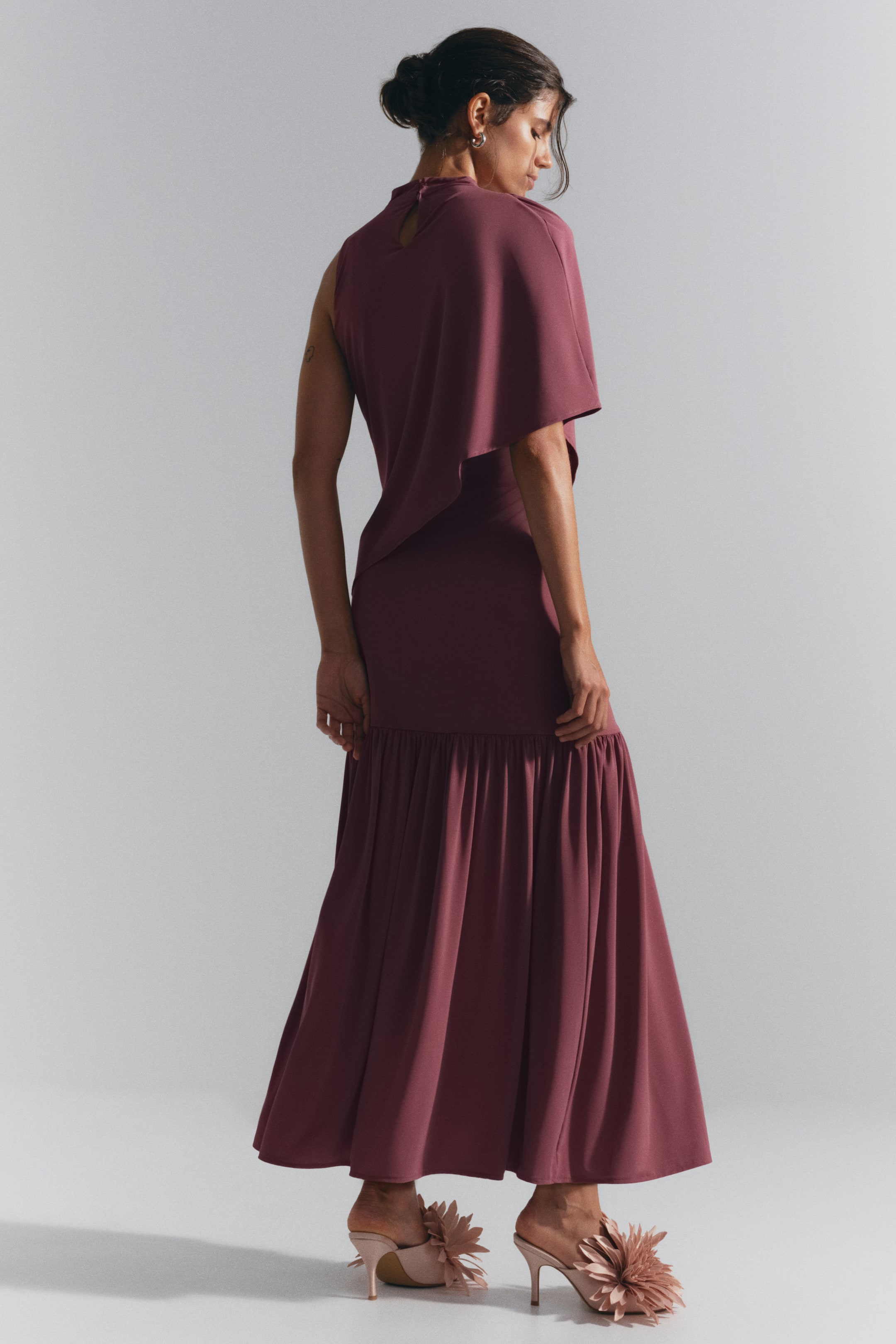 View larger image: A long, sleeveless plum-coloured dress is being worn by the model, shown from a three-quarter back angle. The dress features an asymmetric cape-like layer draped over one shoulder, a keyhole opening at the back of the neck, and a gathered tier below the hip that flows into a wide, flared skirt. The model is styling the dress with light pink, floral-embellished high-heeled mules.