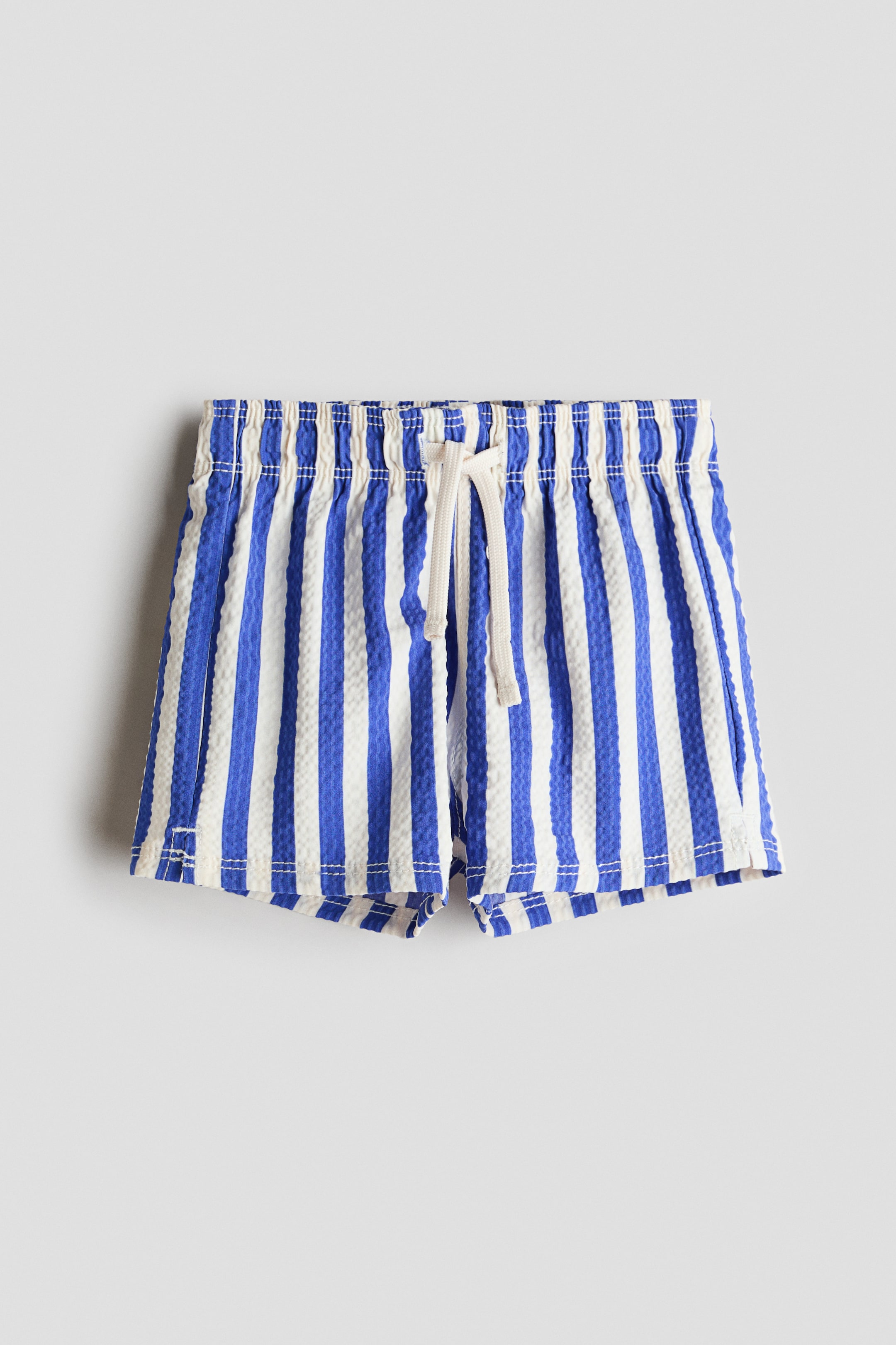 View larger image: These swim shorts showcase blue and off-white vertical stripes on a textured fabric, viewed from the front. They feature an elastic waistband with a visible drawstring and small slits detailing the hems.