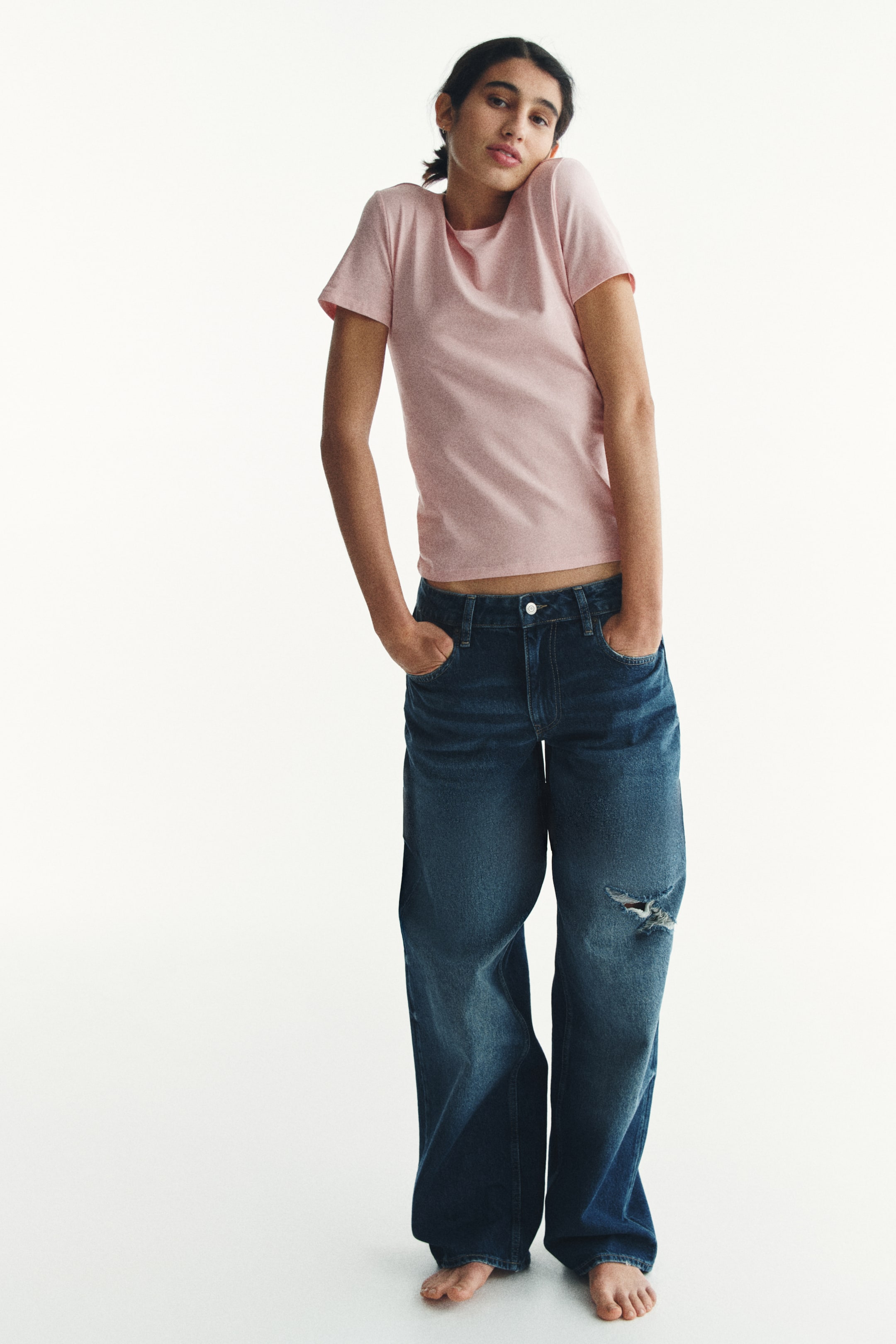 이미지 확대 보기: 연한 핑크색 반소매 티셔츠가 착용되고 있으며, 크루 넥과 허리 위에서 끝나는 크롭 밑단이 특징입니다. 진한 파란색 와이드 레그 청바지도 착용되고 있으며, 오른쪽 무릎에 디스트레스 디테일이 있고, 앞쪽에 버튼 플라이 클로저가 있으며, 손은 앞주머니에 삽입되어 있습니다.