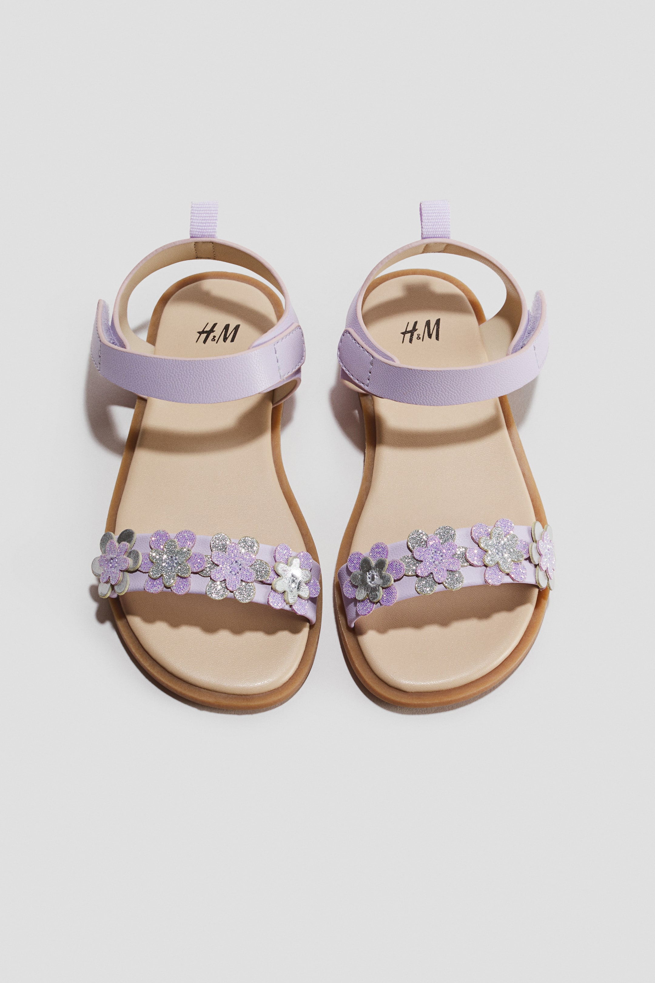 View larger image: A pair of light purple children's sandals is lying flat, showcasing a top-down view of the design. The sandals feature a beige footbed, an adjustable ankle strap with a hook-and-loop closure, and a front toe strap embellished with five small, glittery appliqué flowers in purple and silver.