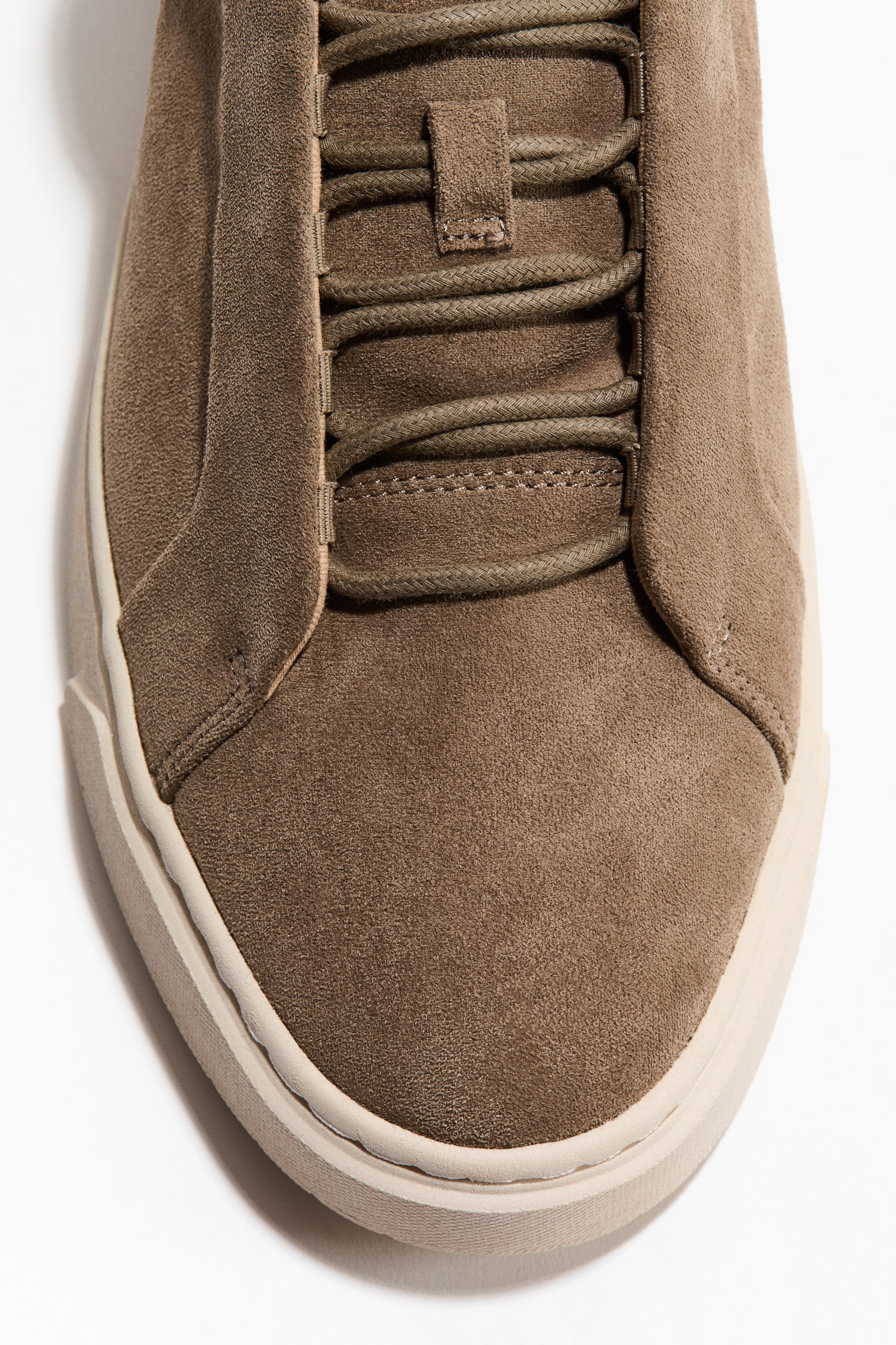 Größeres Bild ansehen: Ein brauner Wildleder-Sneaker zeigt seine Vorderseite, mit einer abgerundeten Spitze und einer hellen Sohle. Passende geflochtene Schnürsenkel fädeln sich durch Ösen, mit einem sichtbaren Wildleder-Schnürsenkelhalter auf der Zunge.