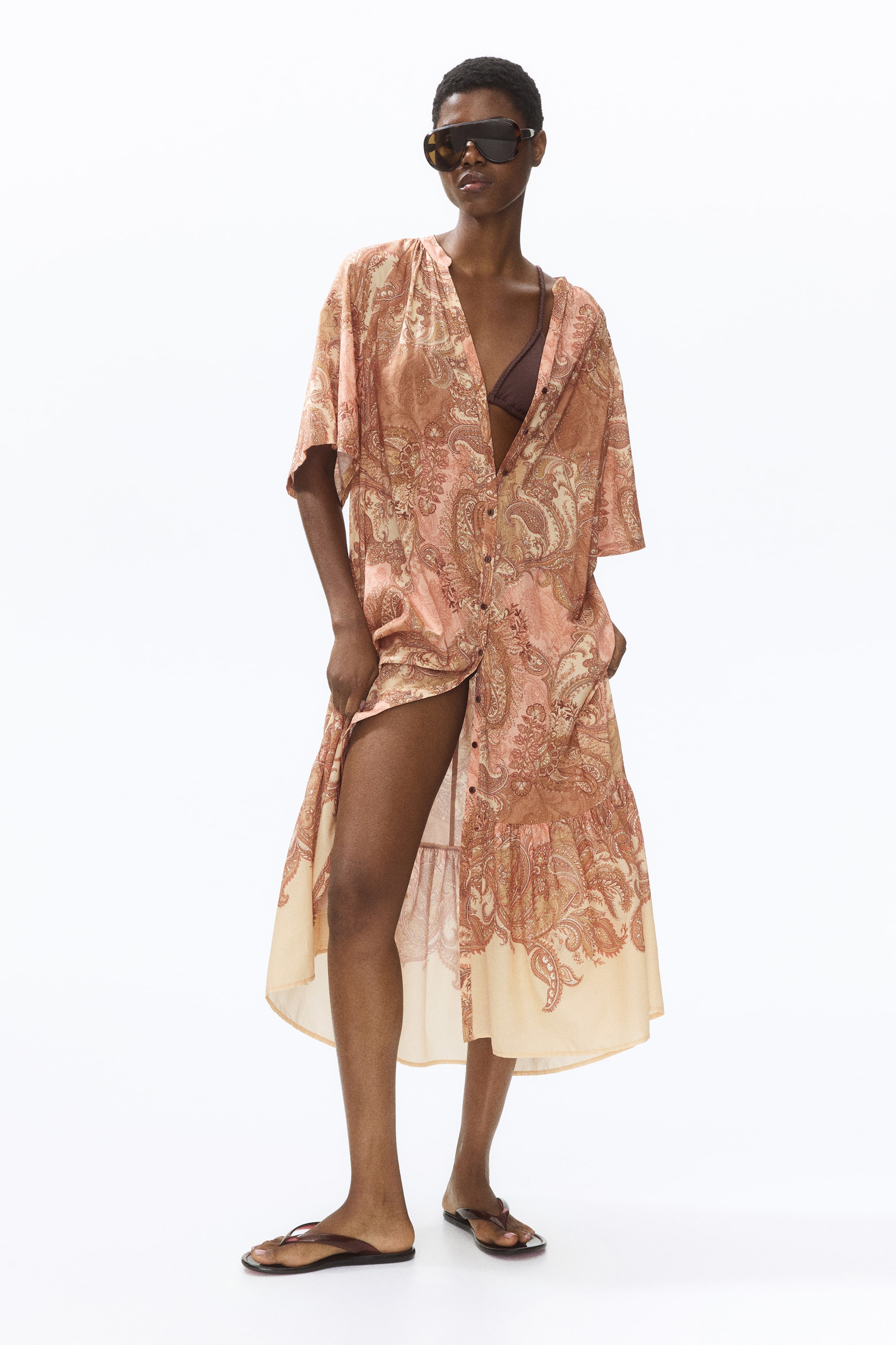 View larger image: A button-front midi dress features a peach and brown paisley print. Designed with short, loose sleeves and a V-neckline formed by the unbuttoned front, it also boasts a tiered ruffle hem with a light solid border. A brown bikini top is visible underneath.