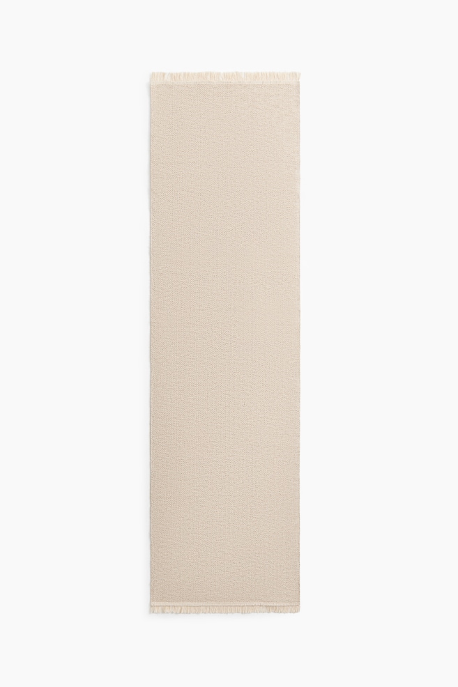A beige woven cotton runner rug is lying flat in a front view, displaying a subtle herringbone pattern and featuring short, light-coloured fringes along its short ends. - 1