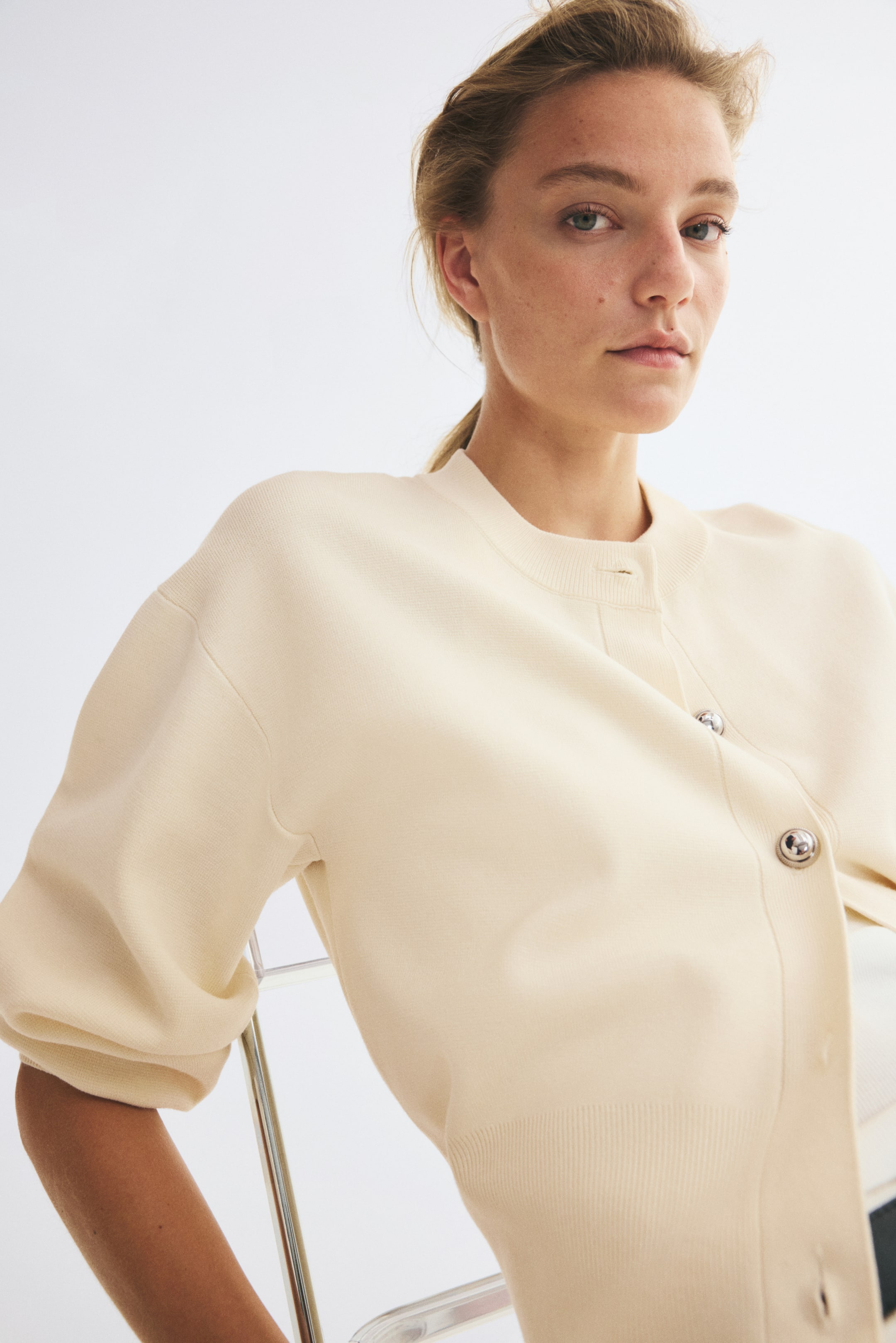 Agrandir l'image: Un cardigan crème en maille fine est présenté de face, orienté vers la gauche, avec un col rond bordé de côtes et des boutons-pression argentés sur le devant. Le modèle présente des épaules tombantes et un ourlet côtelé, avec les manches longues remontées jusqu’aux coudes.