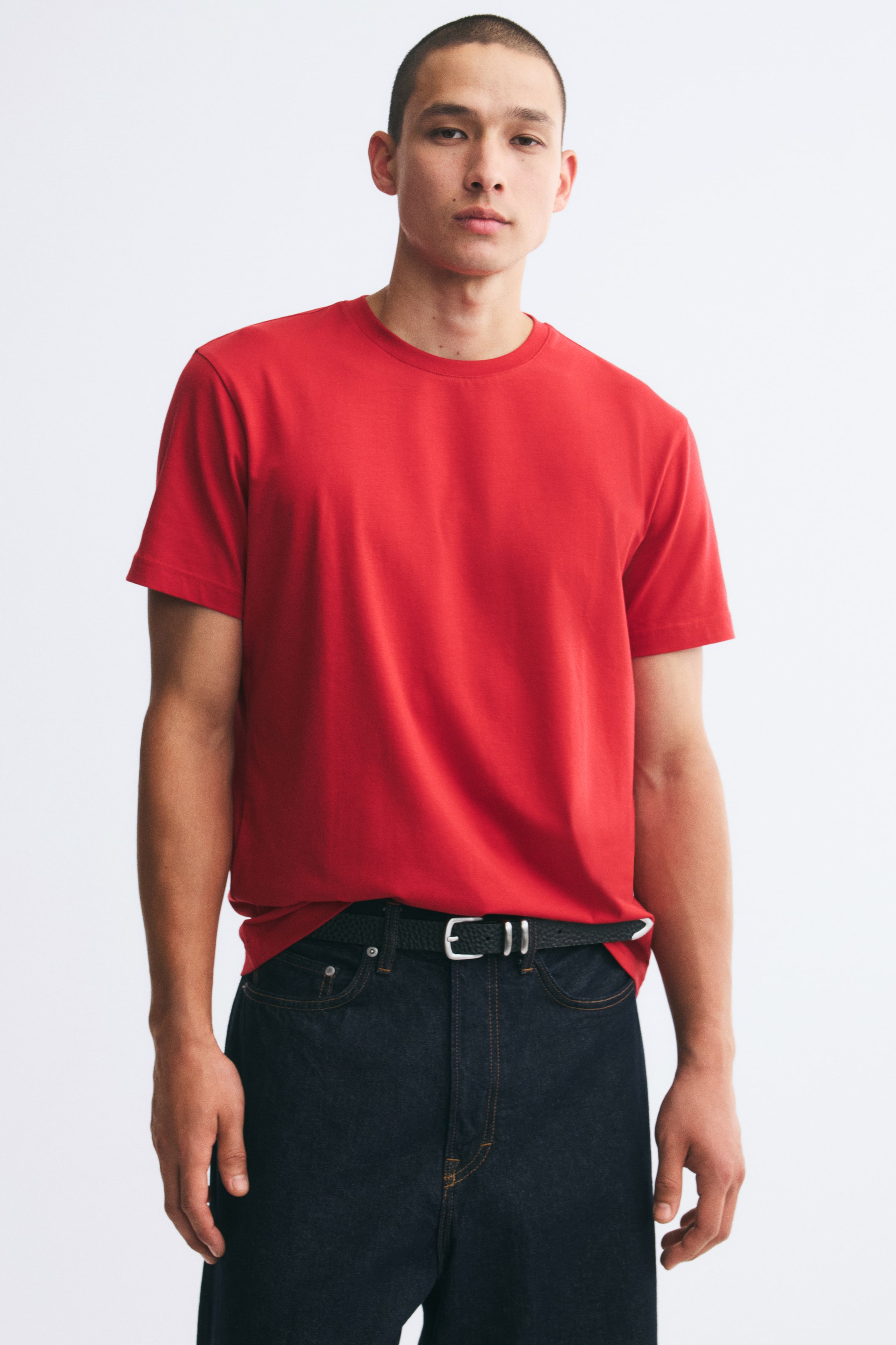 Größeres Bild ansehen: Ein rotes T-Shirt mit kurzen Ärmeln und geripptem Rundhalsausschnitt wird vom Model getragen und betont eine schmale Passform. Das Shirt ist teilweise in die Taille einer dunkelblauen Denim-Jeans gesteckt, die mit einem schwarzen Gürtel kombiniert wird.