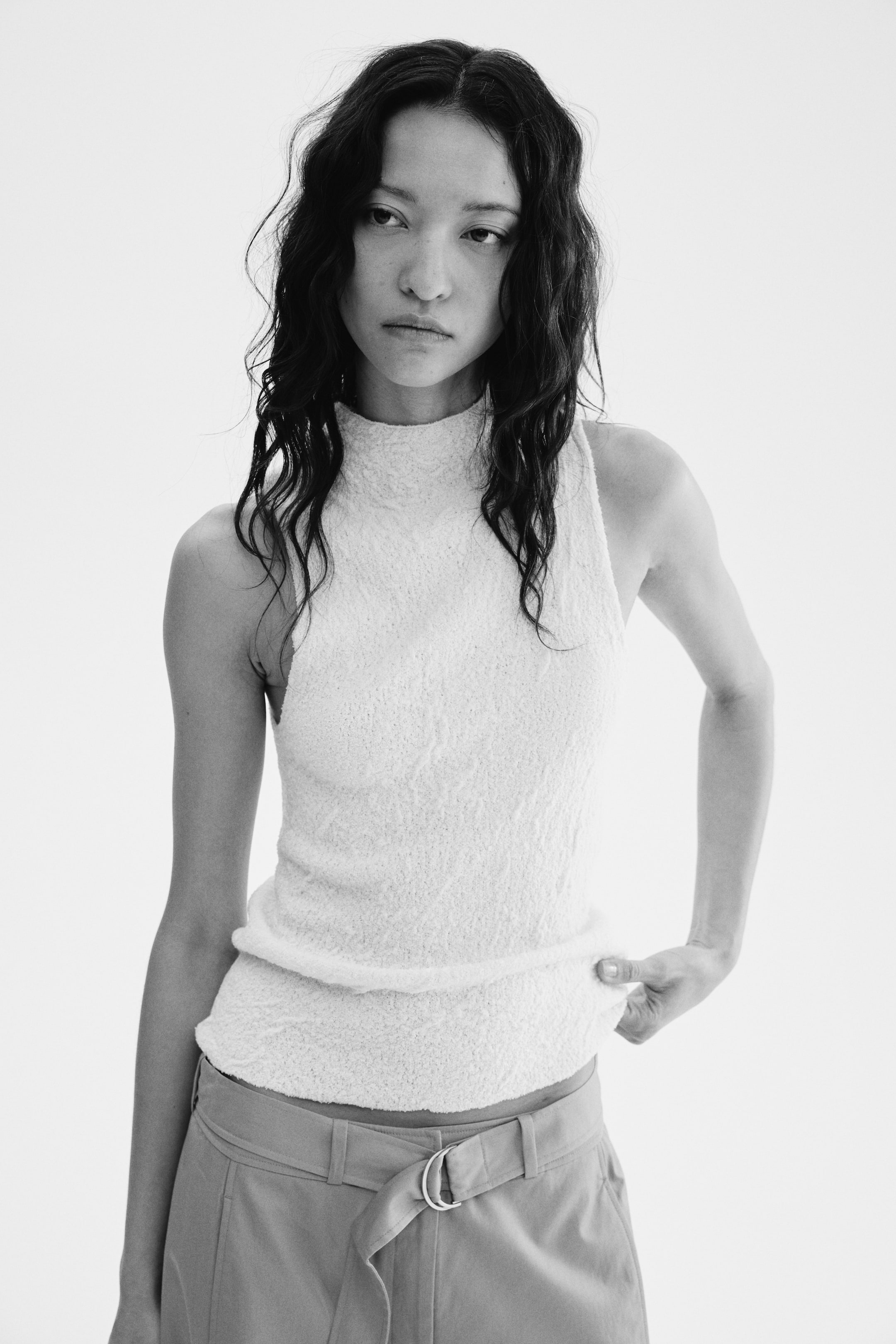 Ver imagem maior: Um top branco sem mangas apresenta uma gola alta e um tecido texturizado e proeminente. O top é apresentado de frente, com a bainha posicionada ligeiramente acima da cintura, sendo ligeiramente puxada para enfatizar o ajuste.