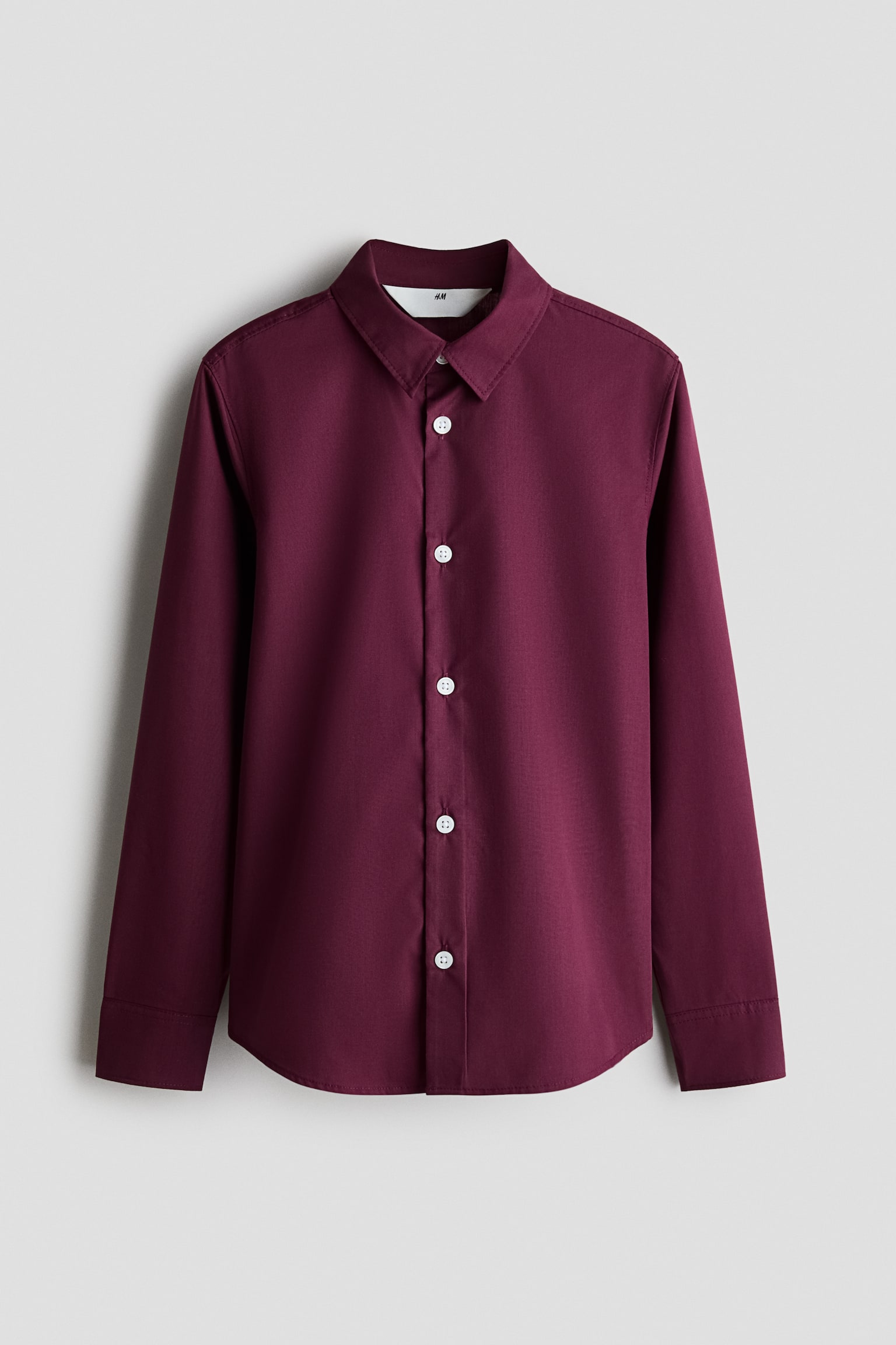 A burgundy long-sleeve poplin shirt is shown from the front, featuring a turn-down collar, white buttons along the front placket, and a rounded hem. The shirt features long sleeves with structured button cuffs.