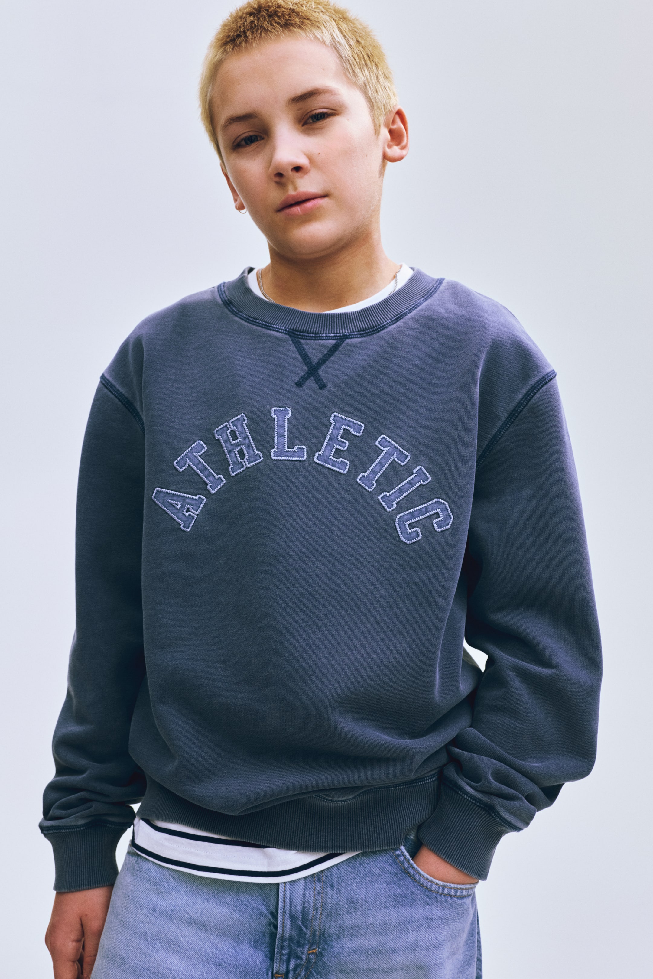 View larger image: Wearing a dark grey crew-neck sweatshirt, featuring "ATHLETIC" in arched, light blue appliqué letters across the chest. Showcasing a washed finish, it includes a ribbed crew neck with a V-stitch detail, and ribbed cuffs and hem, layered over a white striped T-shirt.