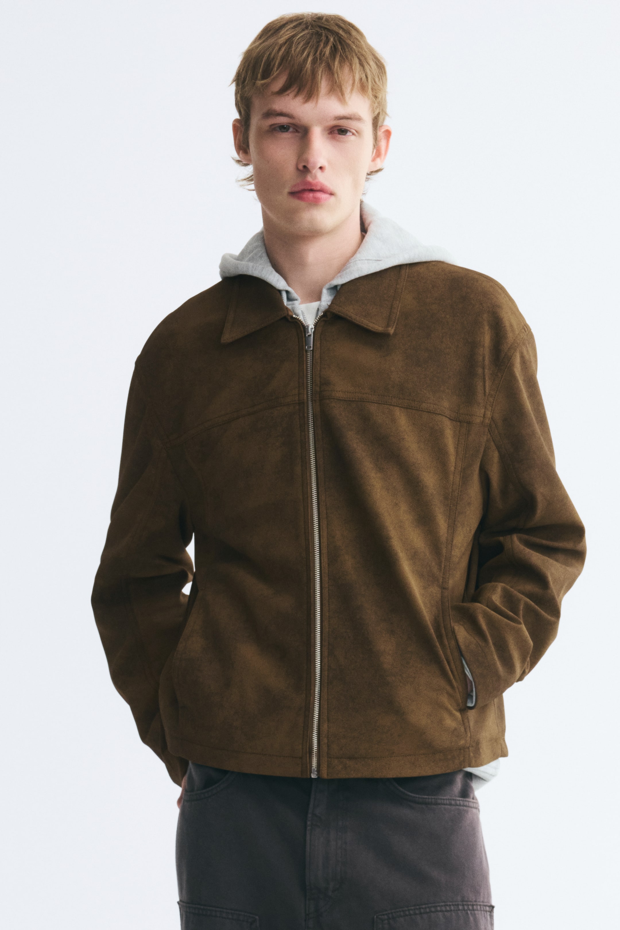 View larger image: A brown napped fabric jacket featuring a collar, a front zip, and a horizontal chest yoke is being worn by the model. The jacket is layered over a light grey hooded sweatshirt, with the model standing with hands tucked into the pockets.
