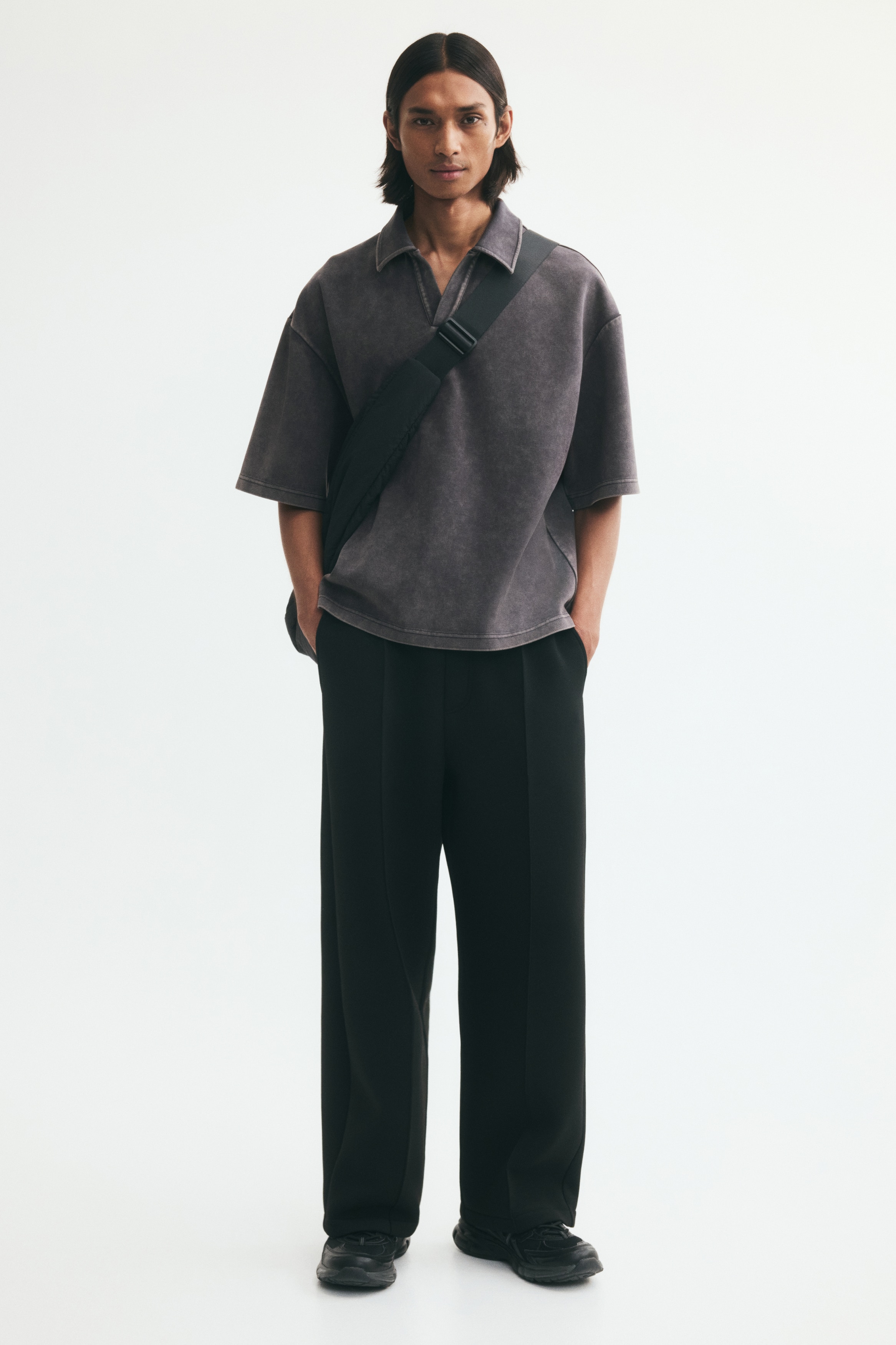 A short-sleeved polo shirt in a washed dark grey finish is facing front, showcasing its relaxed fit and dropped shoulders. It features a flat-lay collar with an open, buttonless V-neck placket and a straight hem.
