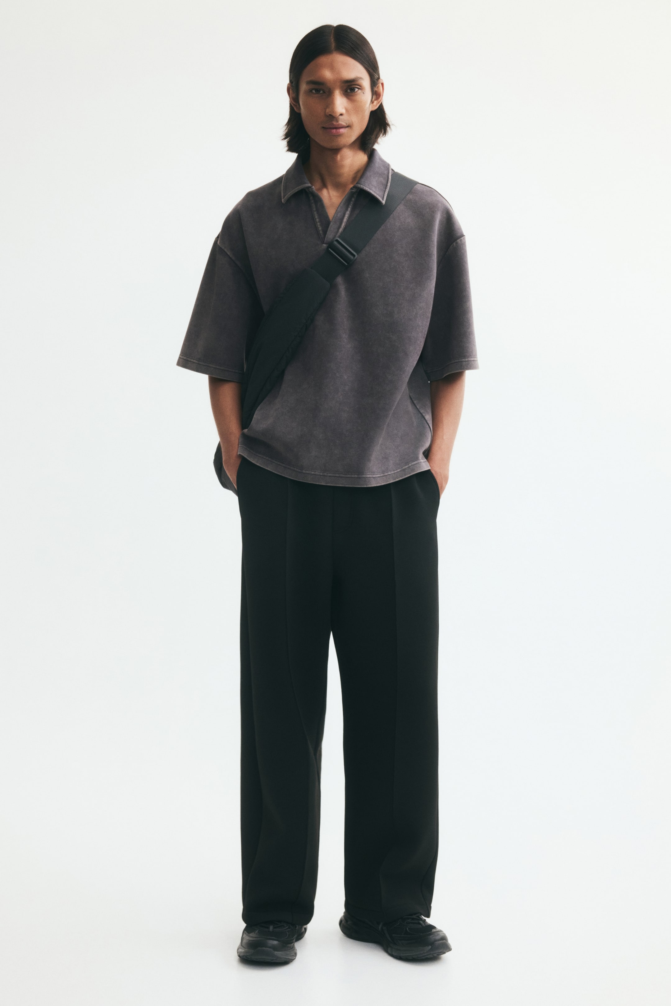 View larger image: A dark gray oversized short-sleeve polo shirt in textured jersey is worn by the model, featuring an open collar, dropped shoulders, and a relaxed silhouette. The shirt is styled with wide-leg black trousers and a black crossbody bag strap worn diagonally across the chest.