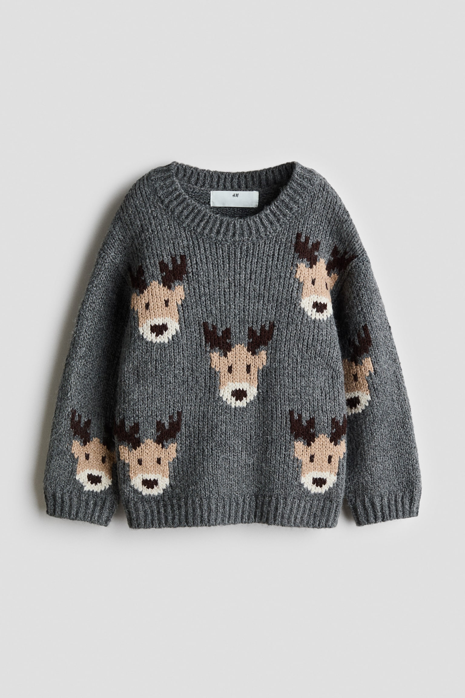 A grey jacquard-knit jumper is shown from the front, featuring a pattern of reindeer faces scattered across the body and sleeves. The jumper is designed with a round, ribbed neckline, dropped shoulders, and ribbing at the cuffs and hem.