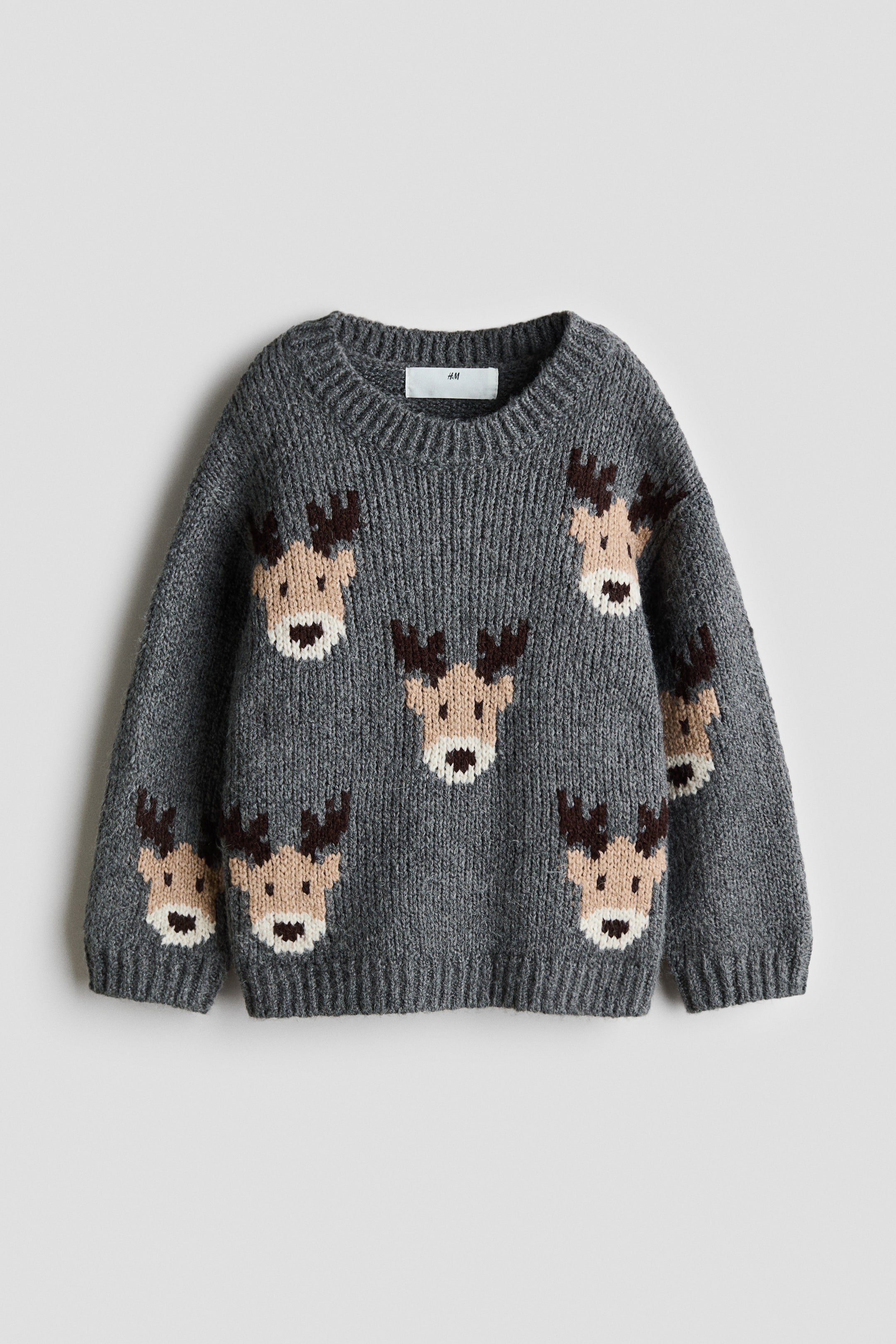 View larger image: A grey jacquard-knit jumper is shown from the front, featuring a pattern of reindeer faces scattered across the body and sleeves. The jumper is designed with a round, ribbed neckline, dropped shoulders, and ribbing at the cuffs and hem.