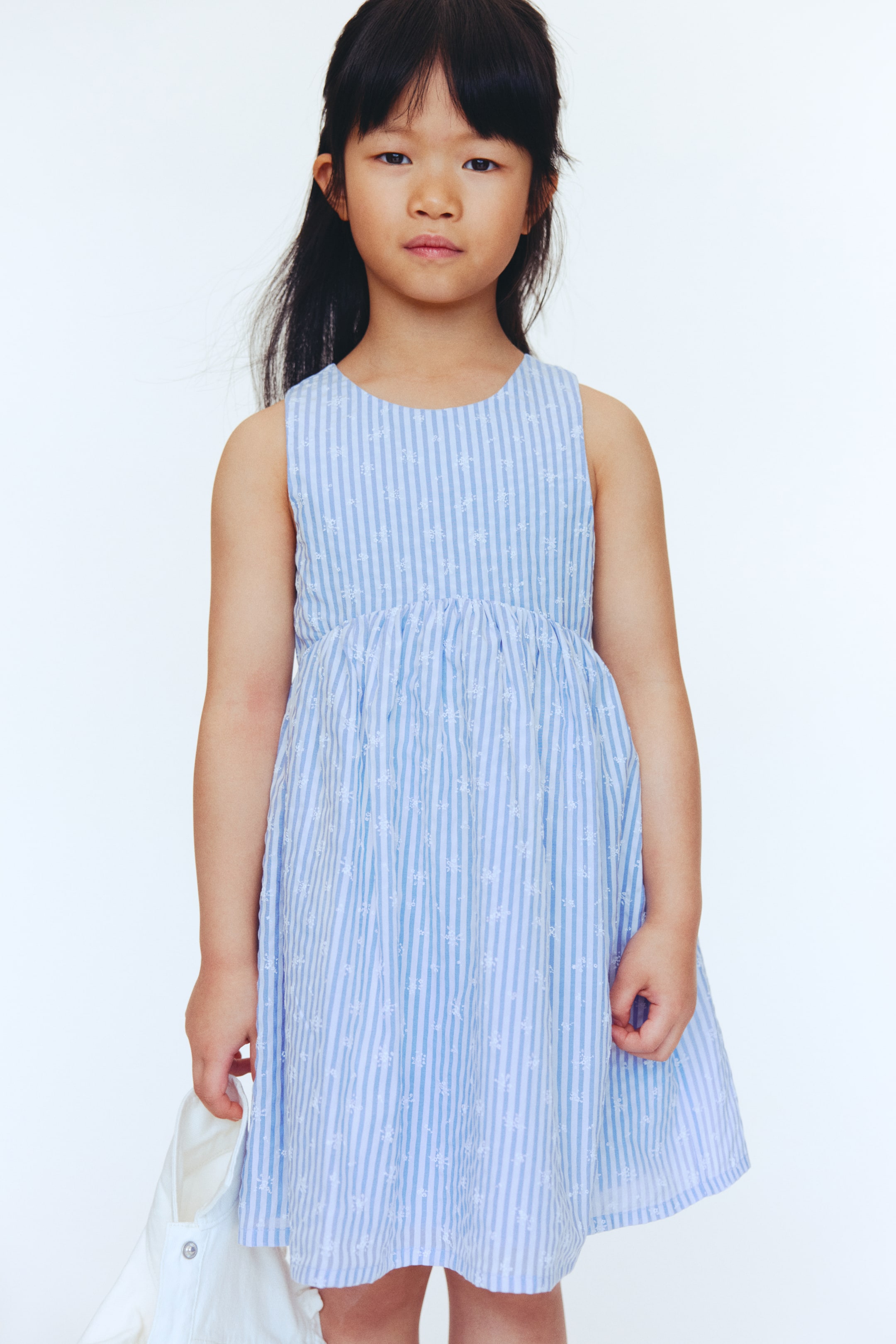 View larger image: A sleeveless light blue and white striped dress is showcased from a front-facing angle. It features a round neckline, an empire waist with a gathered skirt, and white floral embroidery detailing the blue stripes.