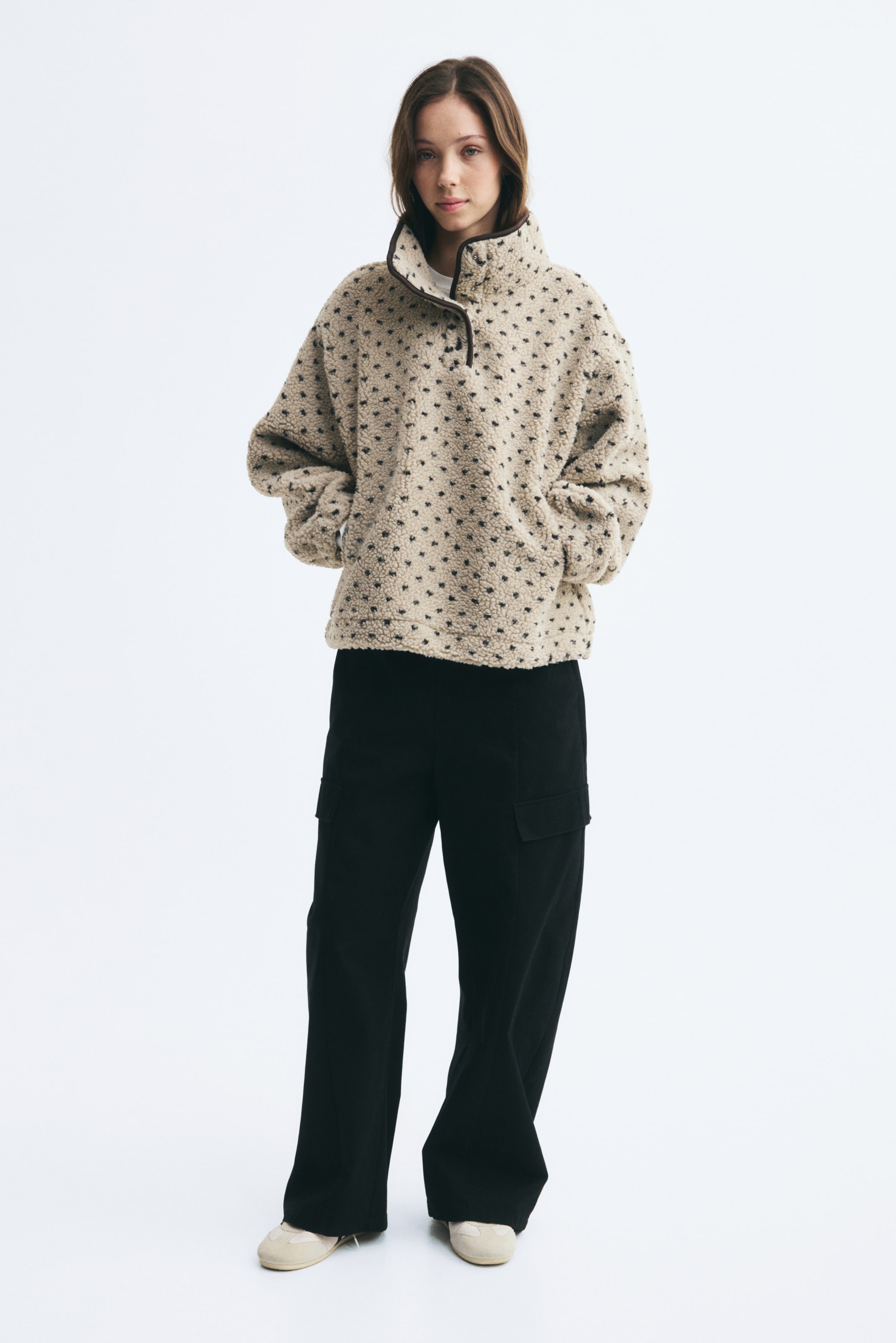 View larger image: A beige teddy fleece sweater featuring a small black printed pattern is worn by the model, who stands with hands placed in the front pockets. The sweater features a high stand collar with dark binding and a partially open button placket, and is styled with black wide-leg trousers.