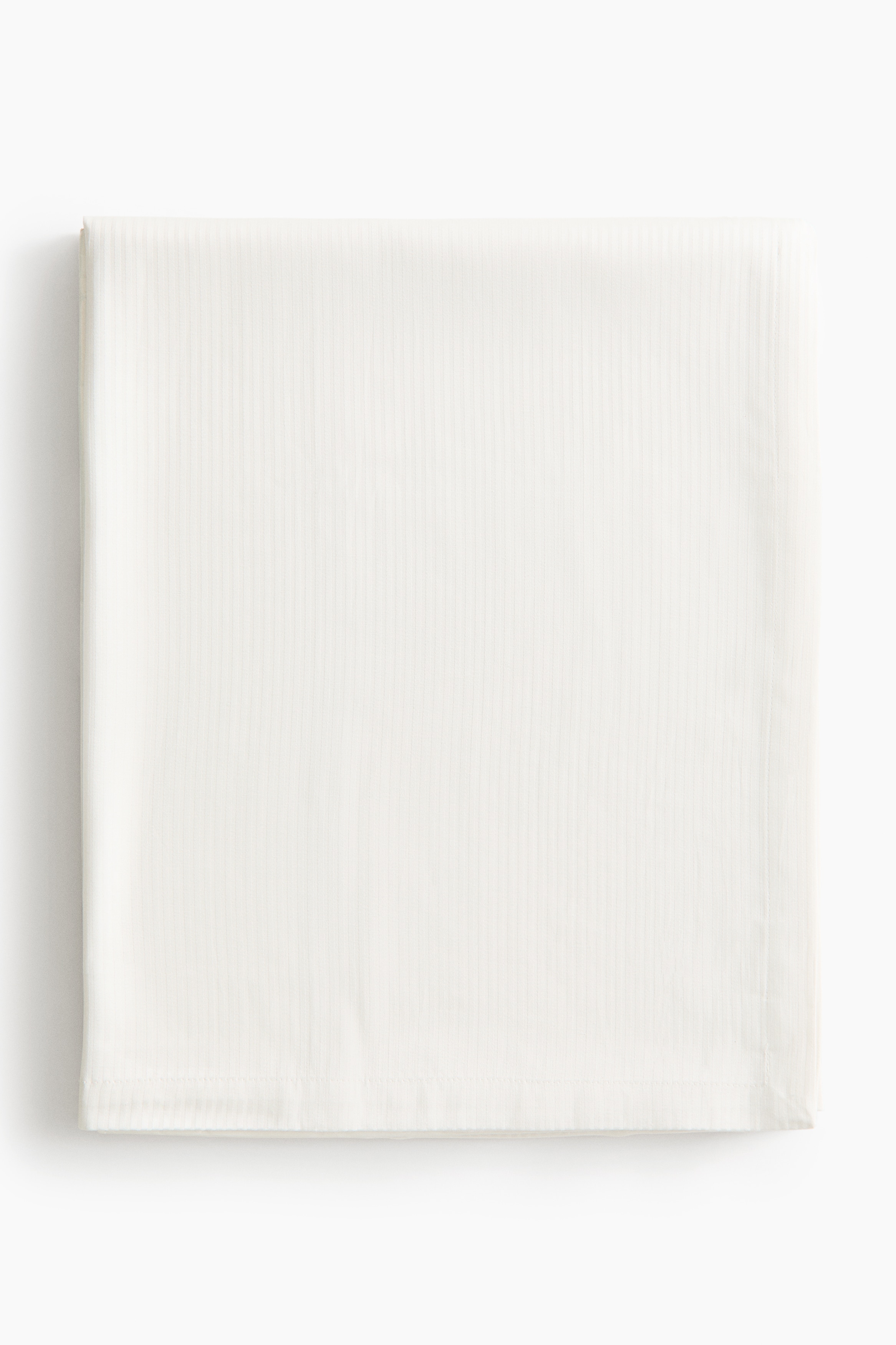 A light beige cotton-blend tablecloth featuring a matching-tone, fine-striped woven pattern is draping over a table corner. The fabric is falling in structured folds, displaying its smooth texture and finished folded hem.
