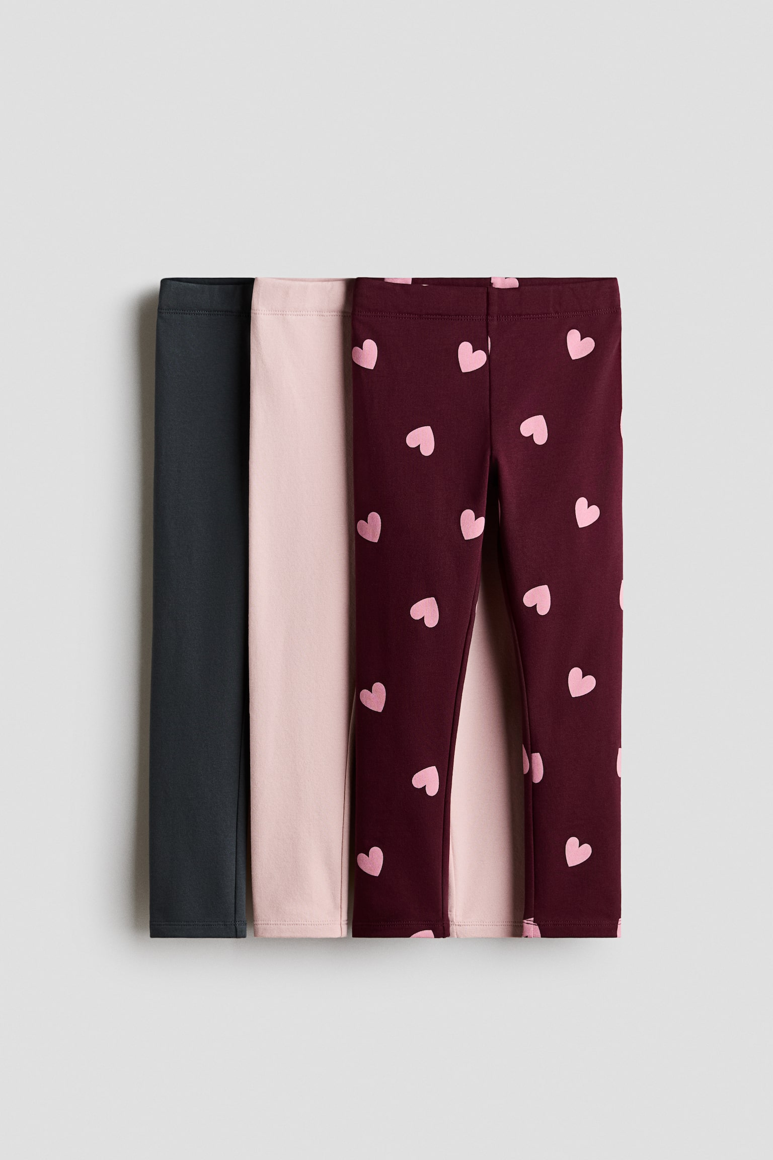 Three pairs of leggings are overlapping from the front to showcase their designs, featuring elasticated waistbands. The set includes a solid dark grey pair, a solid light pink pair, and a burgundy pair with an all-over pattern of small light pink hearts.