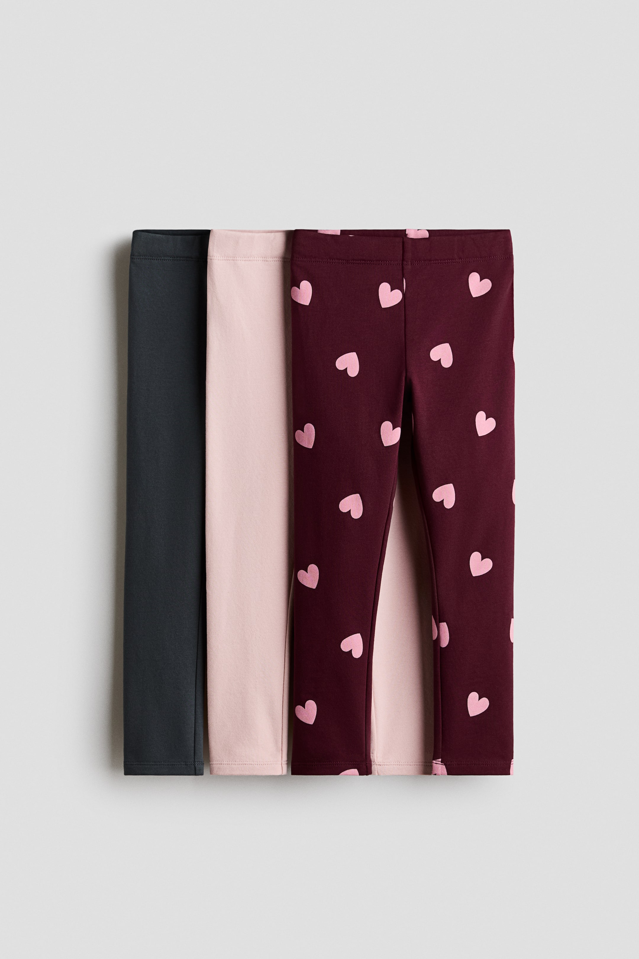View larger image: Three pairs of leggings are overlapping from the front to showcase their designs, featuring elasticated waistbands. The set includes a solid dark grey pair, a solid light pink pair, and a burgundy pair with an all-over pattern of small light pink hearts.