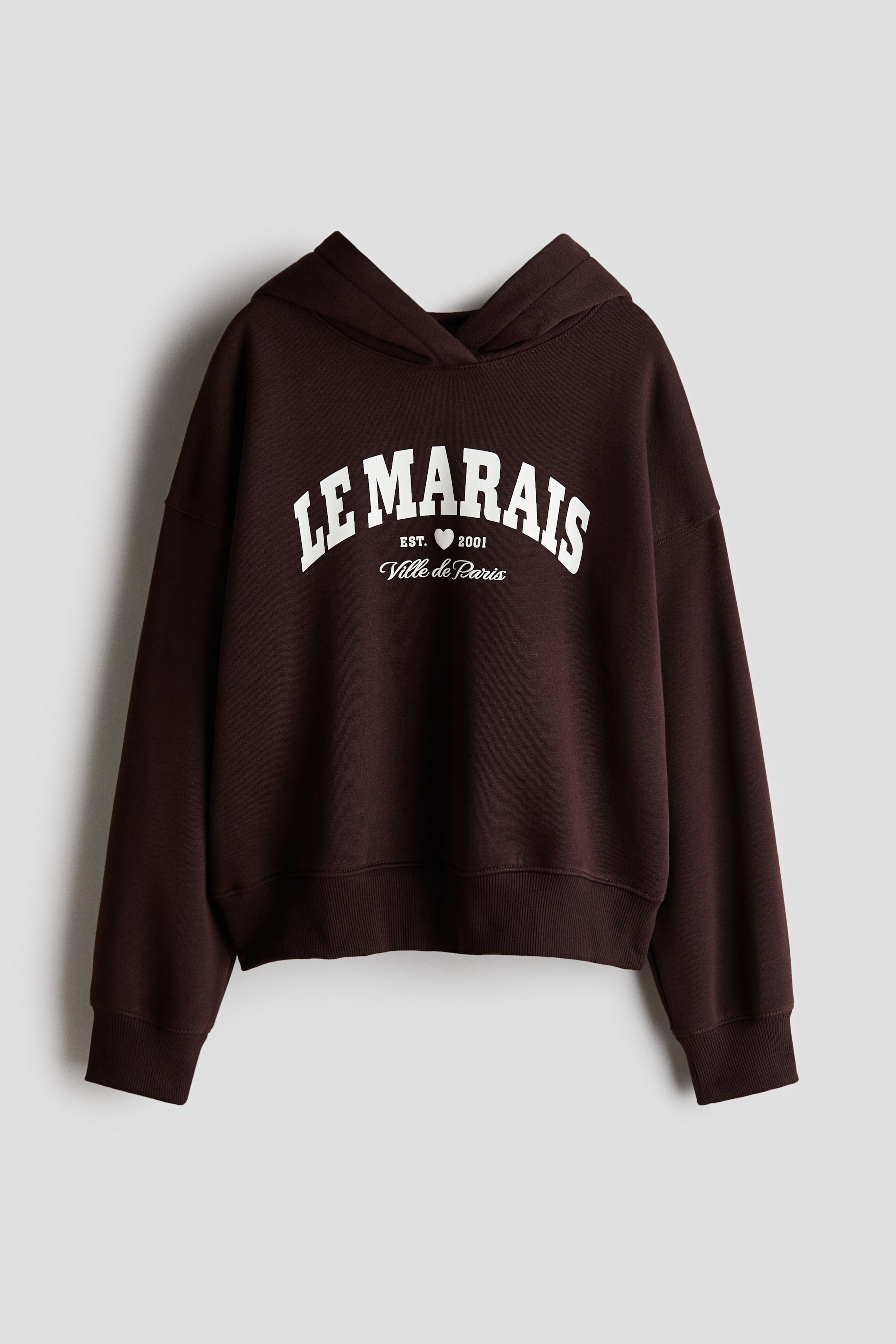 View larger image: A dark brown fleece hoodie is shown from the front, featuring a crossover wrap hood, dropped shoulders, and long sleeves with ribbed cuffs and hem. The chest is accented with a white varsity-style graphic print reading "LE MARAIS," with "EST. 2001" and "Ville de Paris" underneath.